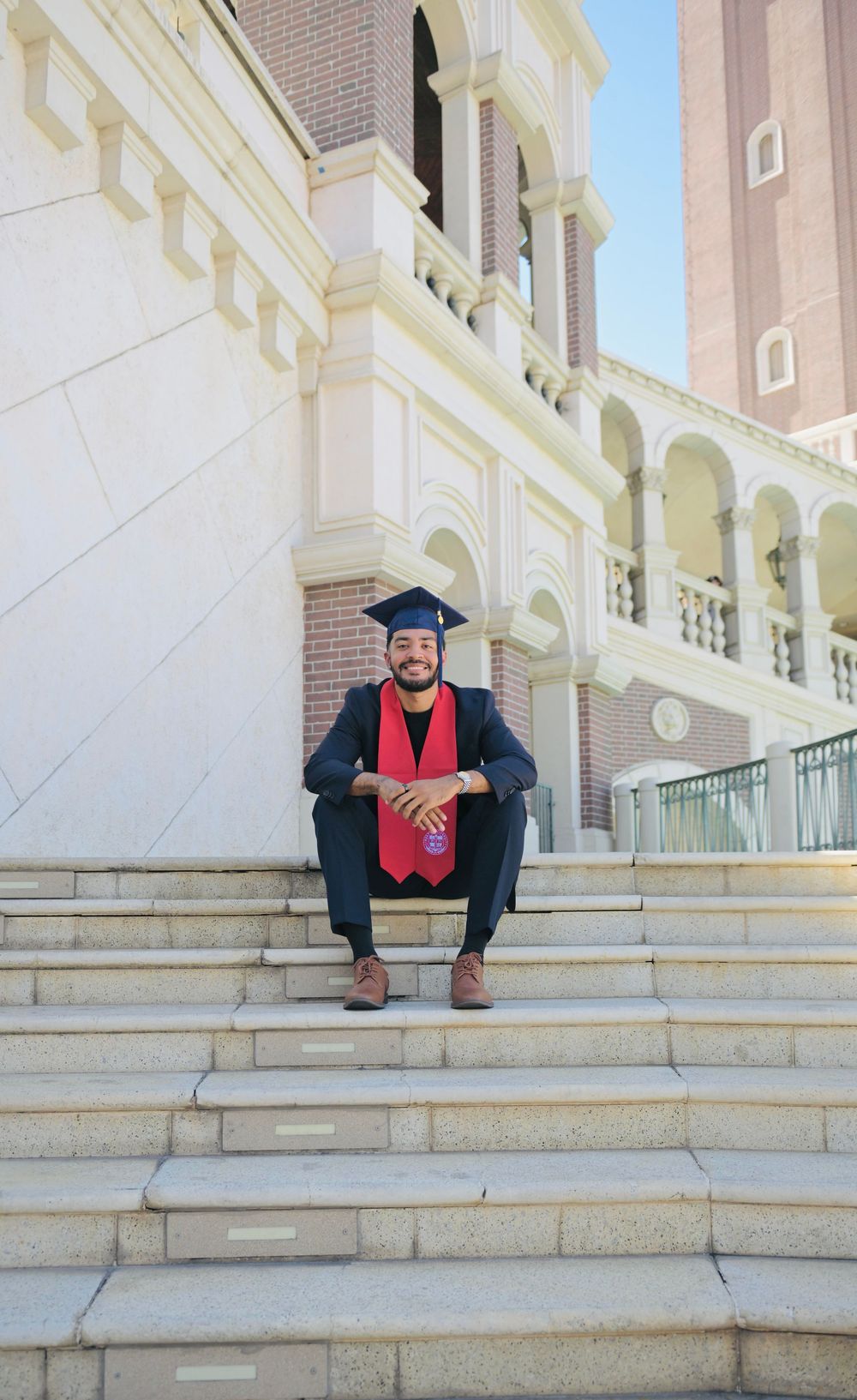 A smiling graduate in cap and gown sits on university steps wearing a red stole, posing for graduation photos.