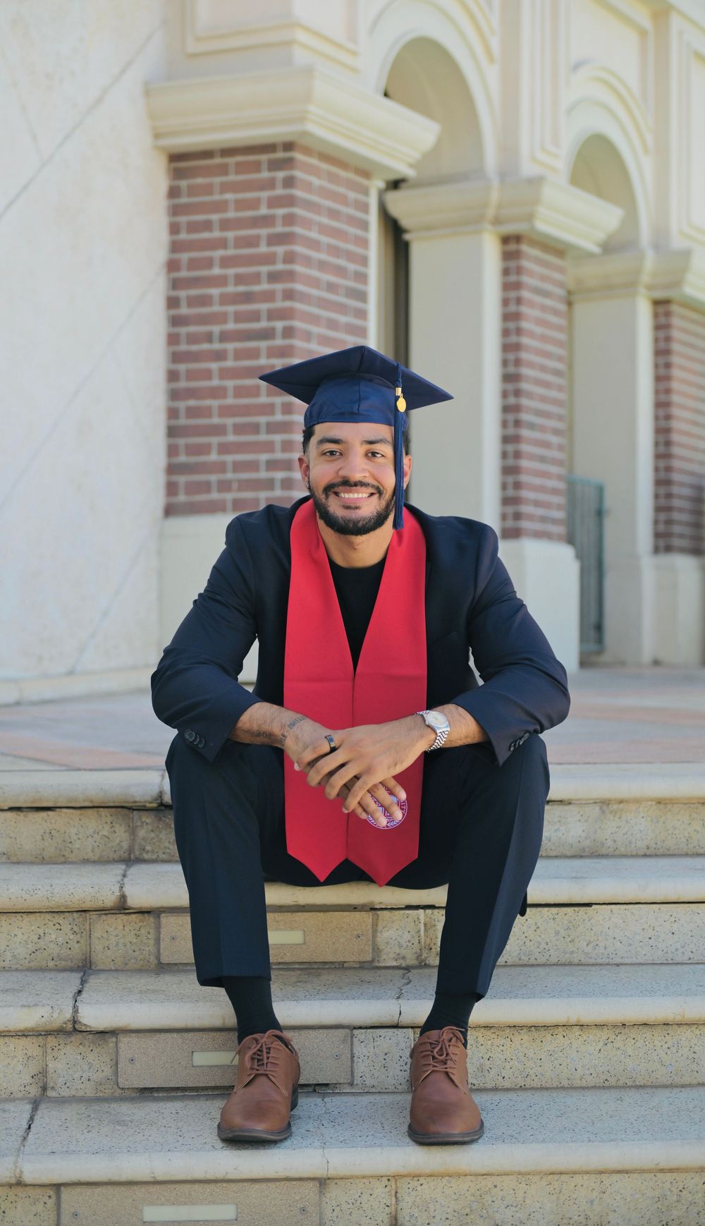 Smiling male graduate in cap and red stole sits on steps in navy suit outside brick building.