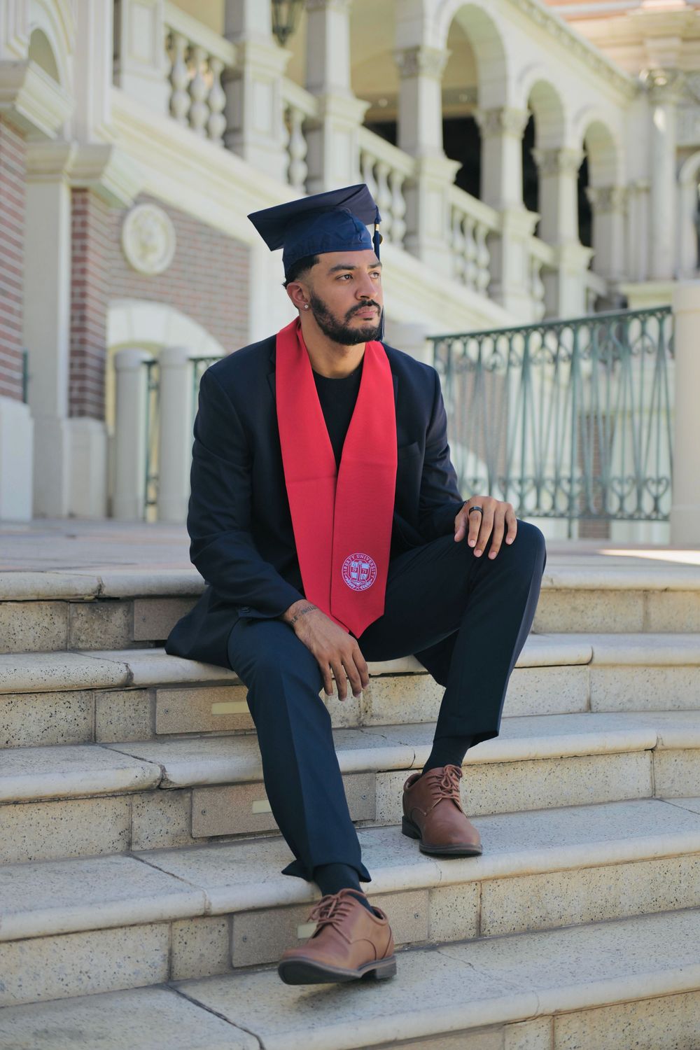 Graduate in cap, red stole, and navy suit sits on stone steps outside a classical building.