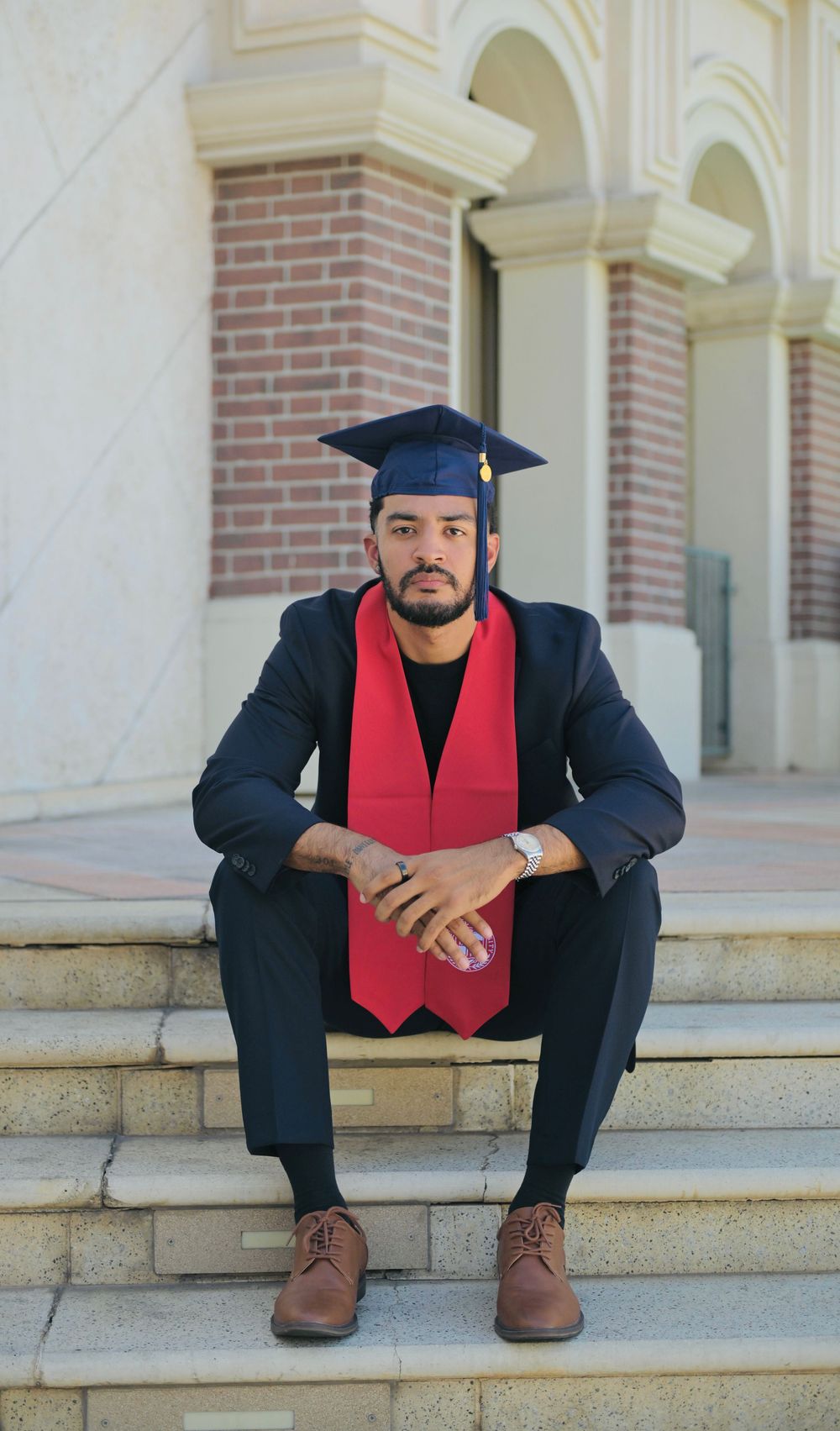 A graduate in cap and red stole sits on university steps in a dark suit and brown shoes.