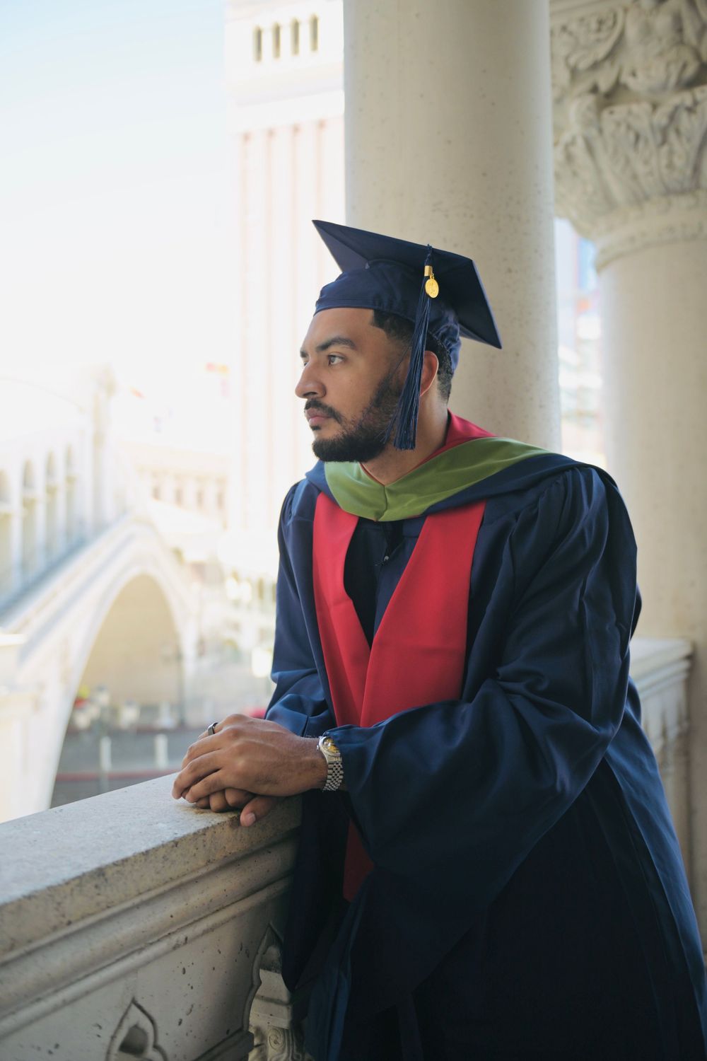 Young man in navy graduation gown with green hood and mortarboard cap leaning on balcony railing.