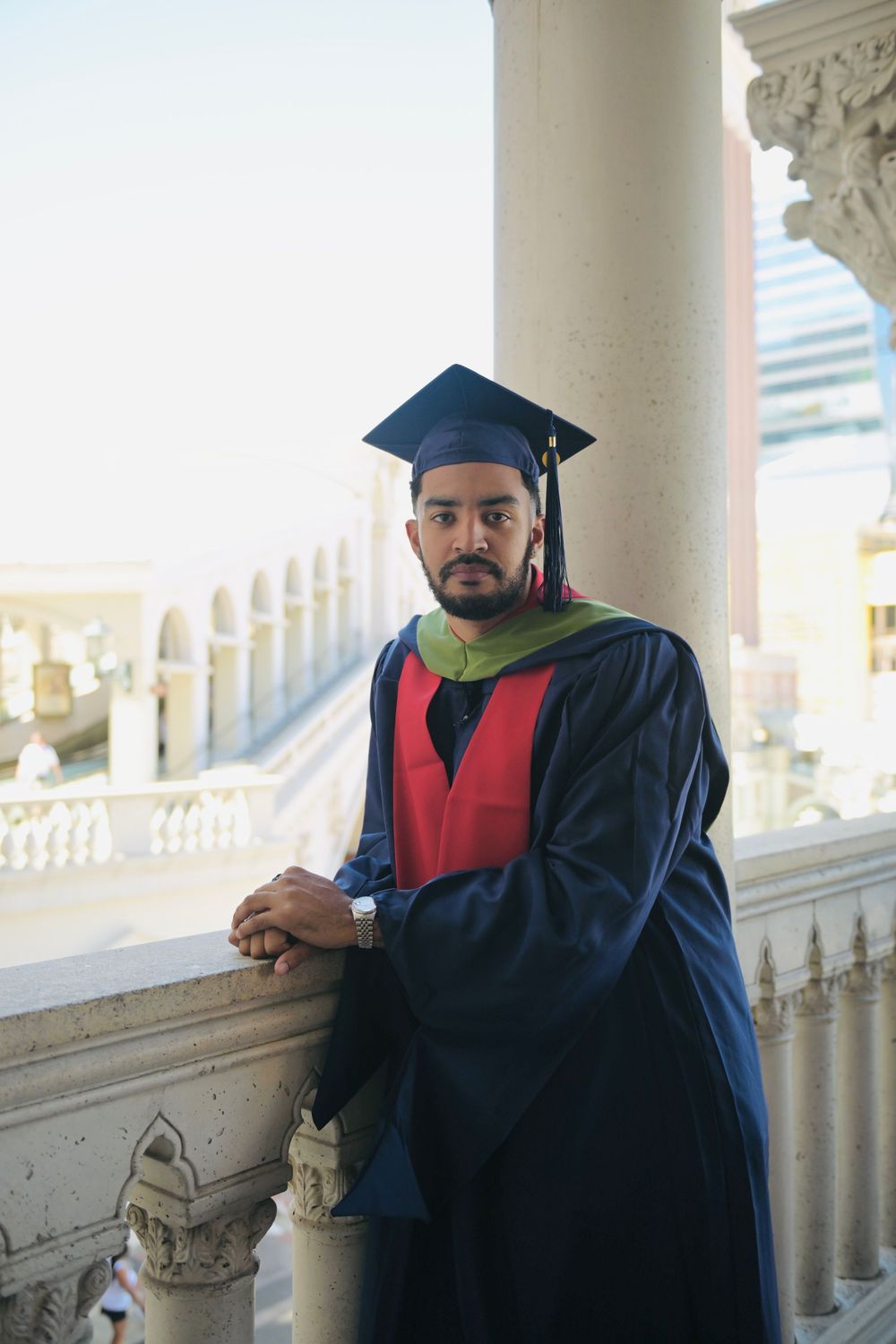 A young man in graduation cap and gown poses on an ornate balcony, wearing a blue robe with red and green accents.