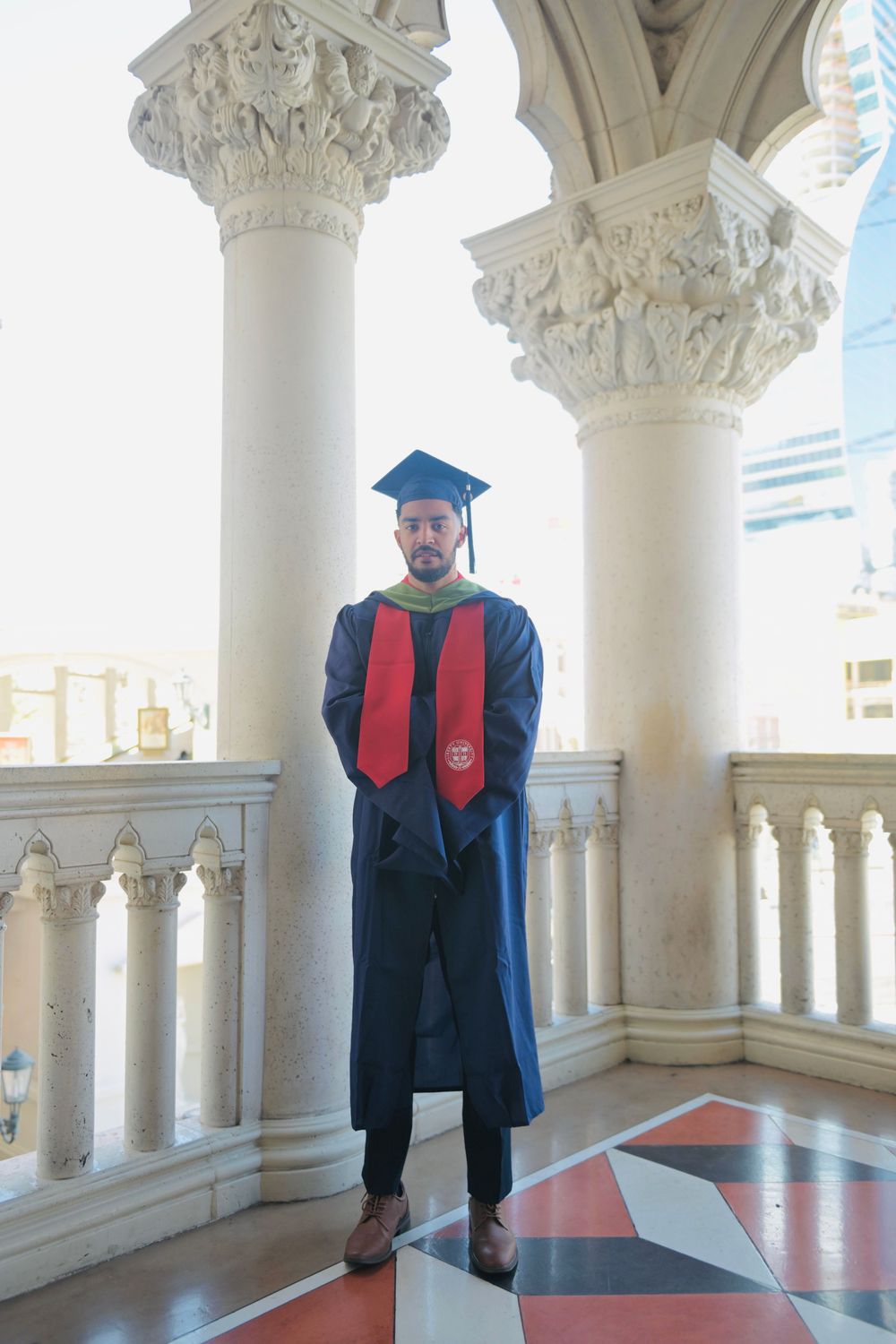 Graduate in cap and gown with red stole poses between ornate white columns at an elegant venue.