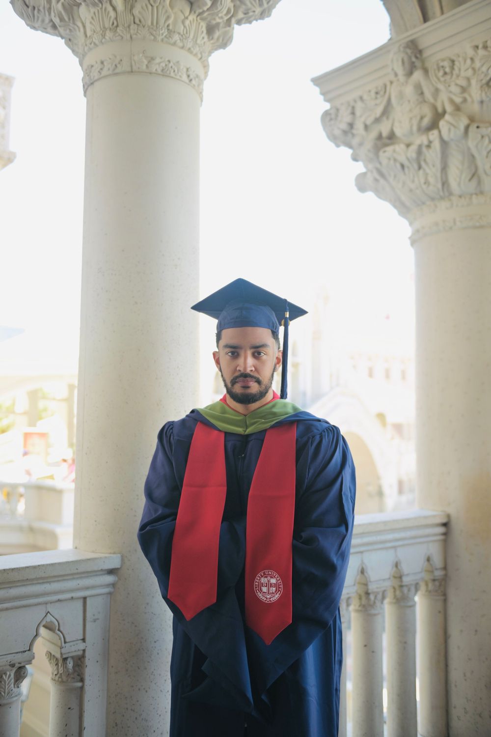 Graduate in blue cap and gown with red and green stoles poses between white columns at a university.