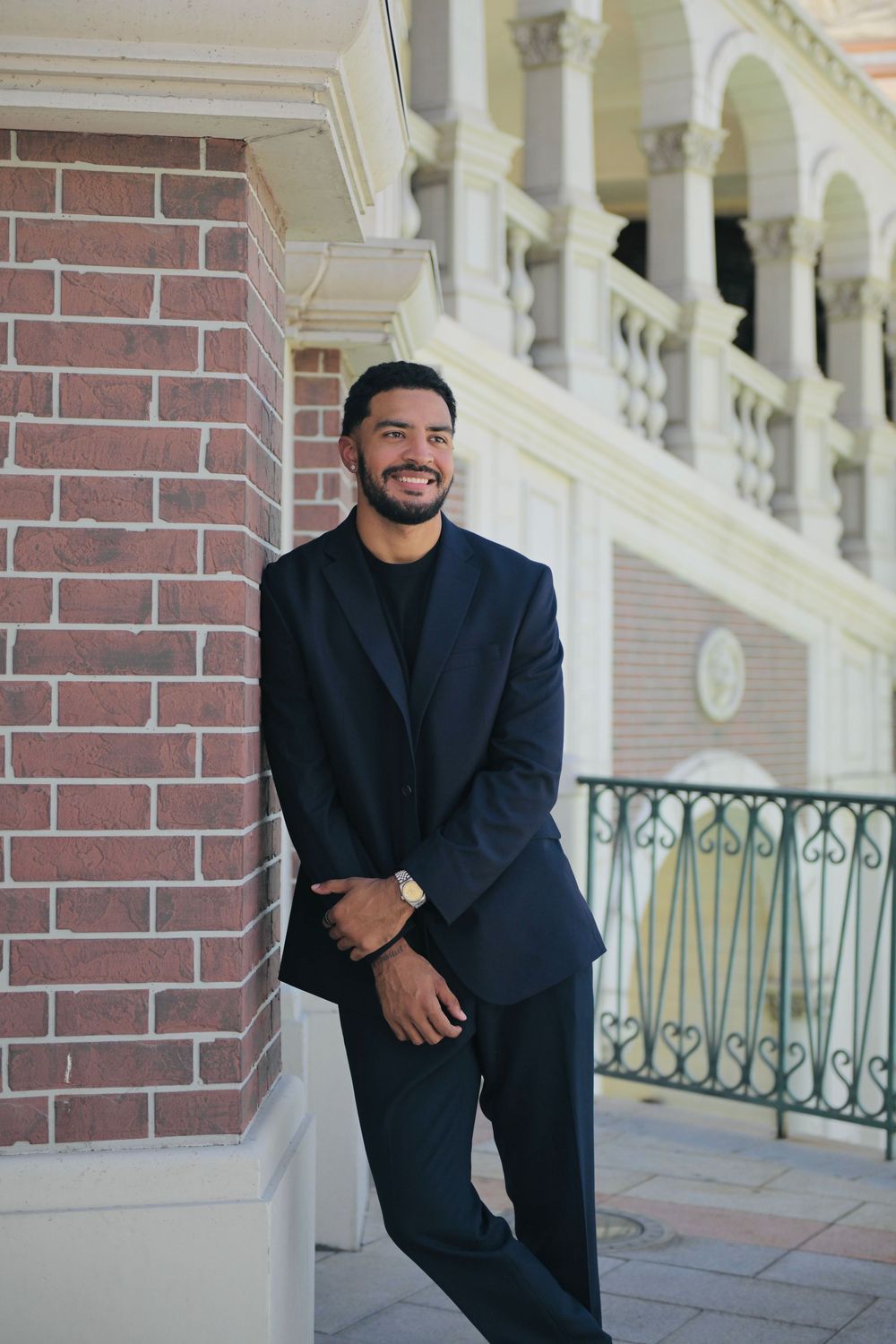 Smiling young man in a navy suit leaning against a brick column at an elegant building with arched architecture.