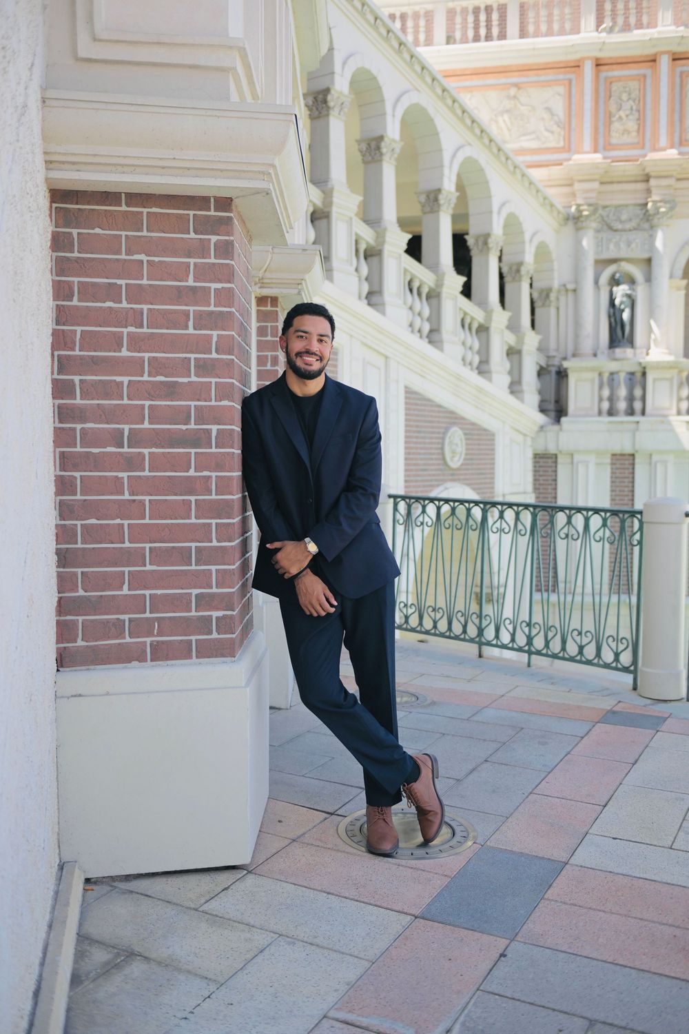 Smiling man in navy suit leaning against brick column at elegant building.