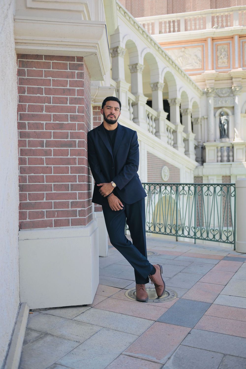 Man in navy suit leaning against brick wall at ornate building.
