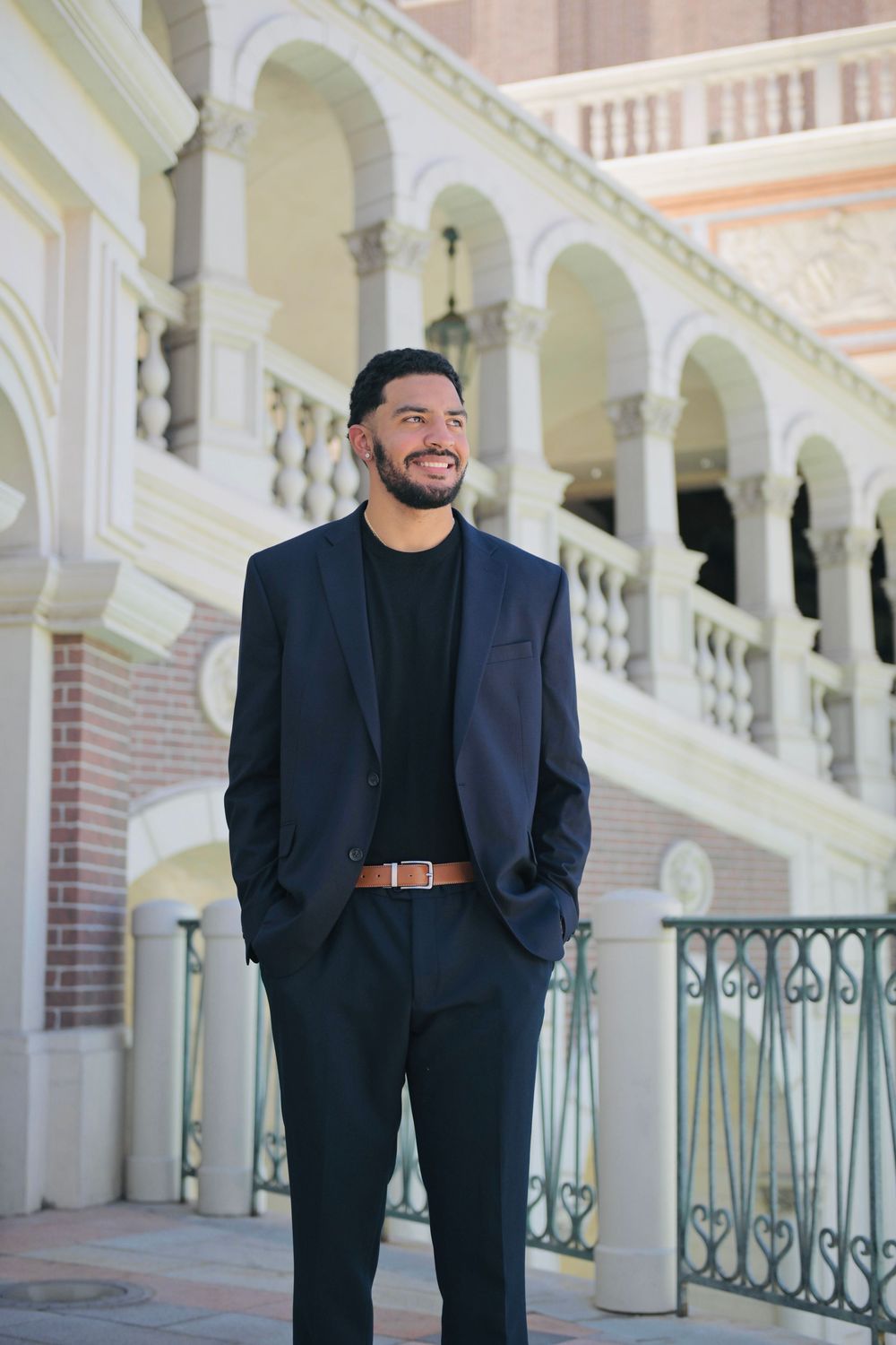 Smiling man in navy suit and black shirt stands confidently outdoors near elegant arched building with ornate railings.