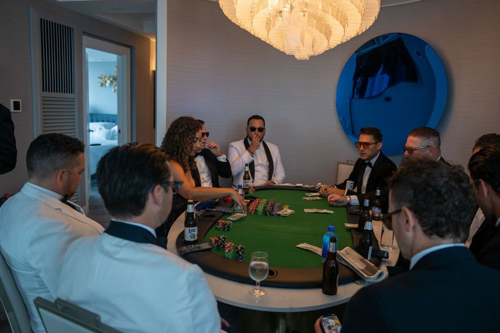 Group of elegantly dressed people playing poker around a green felt table in a modern room with chandelier.
