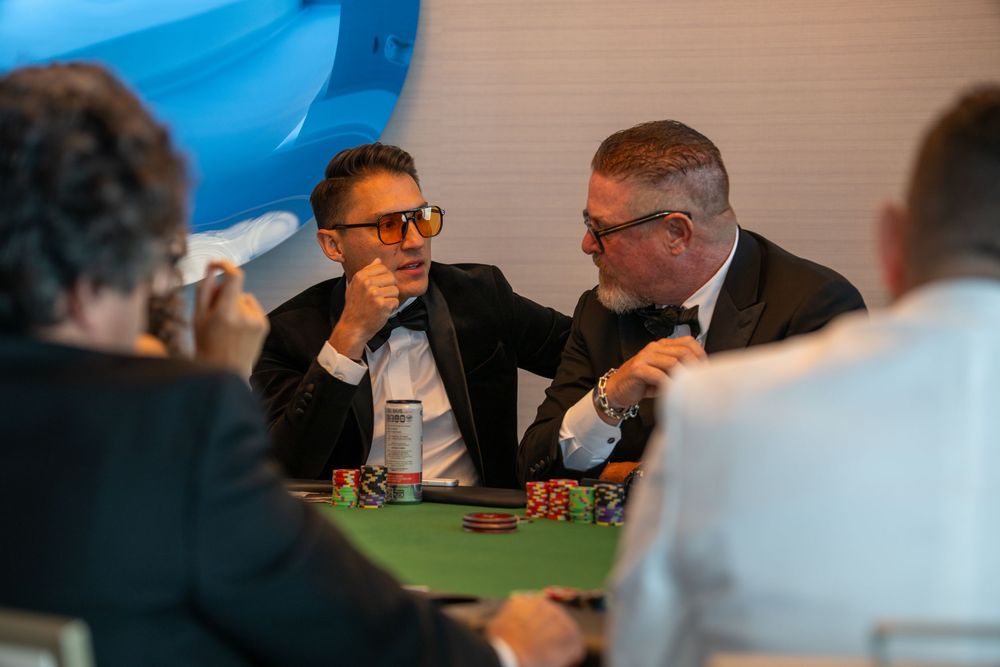 Two men in suits playing poker at a green felt casino table with colorful chips stacked during a formal event.