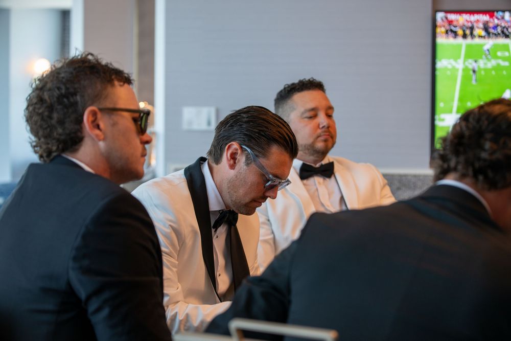 Men in formal attire gathered around a table at an event, with a football game on TV in the background.