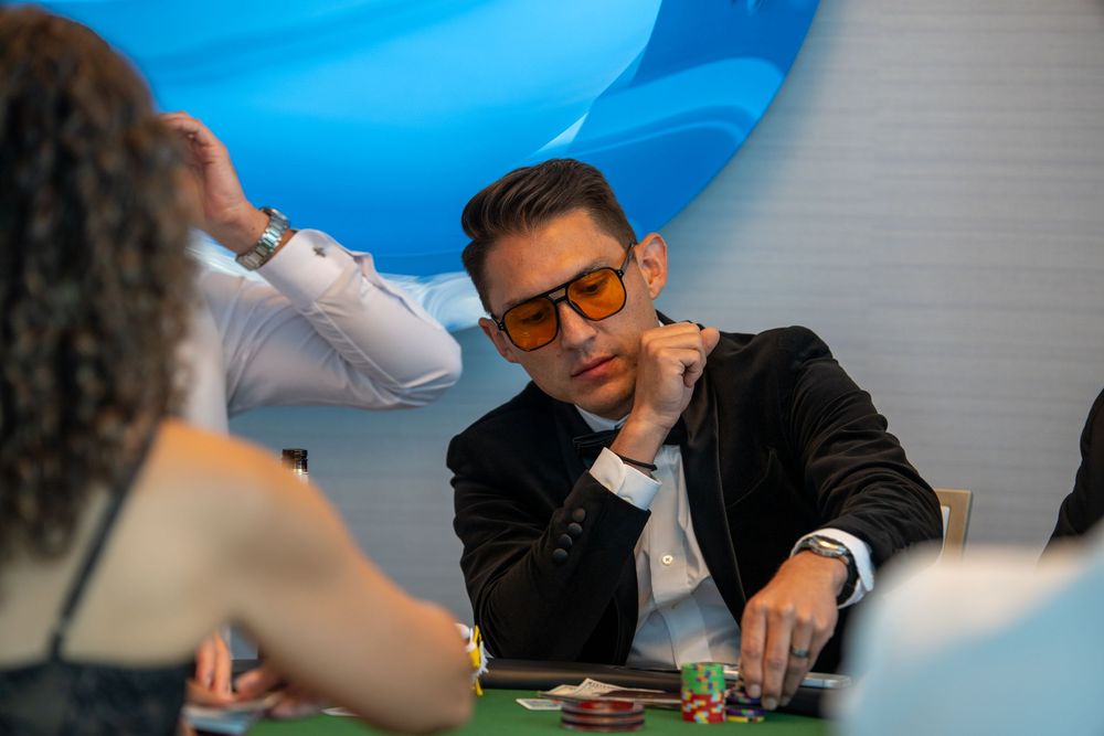 Man in sunglasses and black blazer focuses intently at a poker table during a card game.