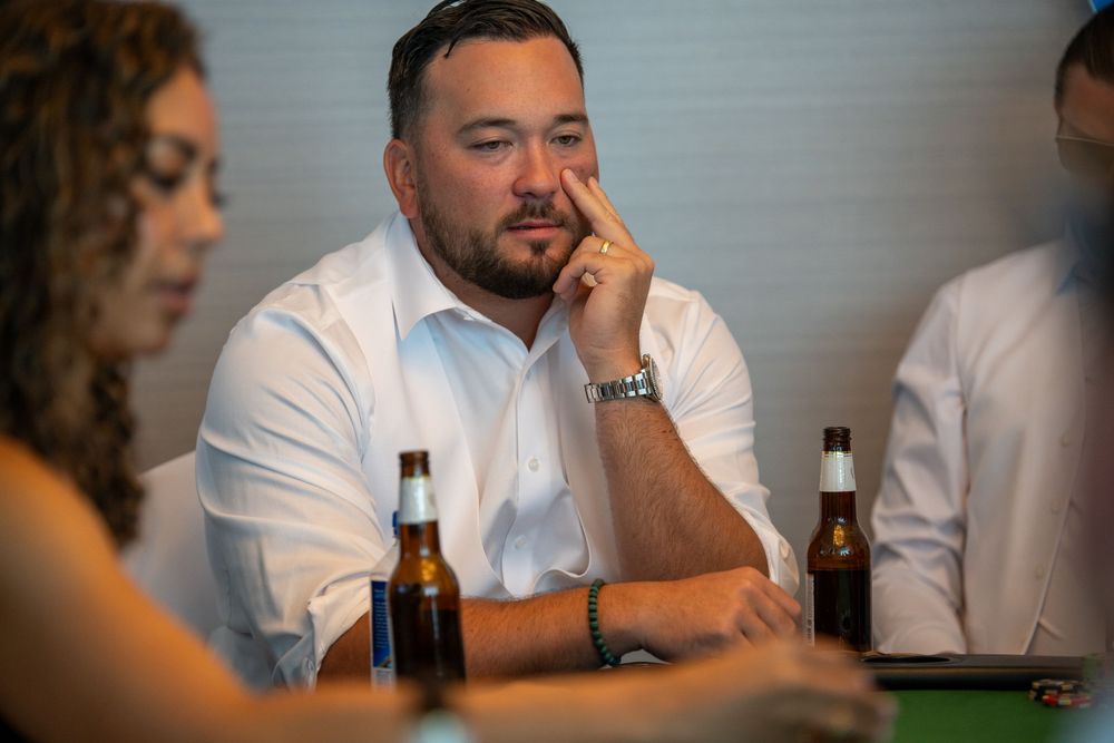 Man in white shirt sitting at table with beer bottles, looking thoughtful during a casual social gathering.