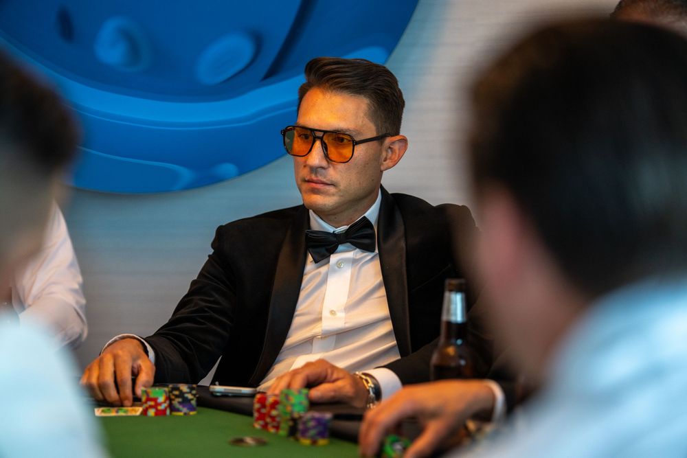 Man in tuxedo wearing orange sunglasses playing poker at a casino table with poker chips.