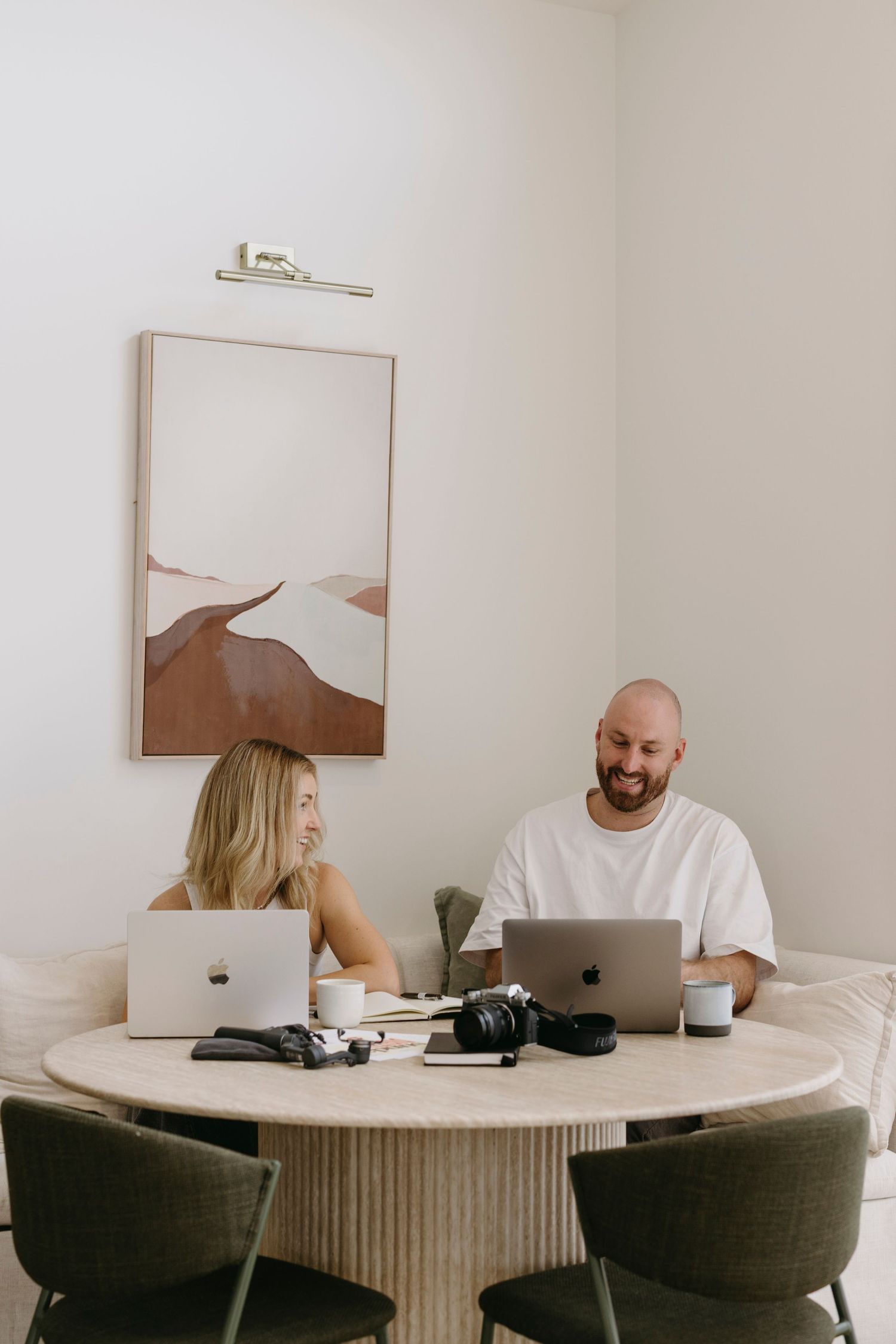 A smiling man and woman work on laptops at a round wooden table in a modern, bright home office.