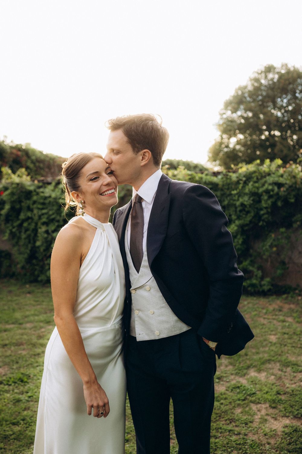 Groom in dark suit kisses bride on cheek as she smiles in white halter dress outdoors.