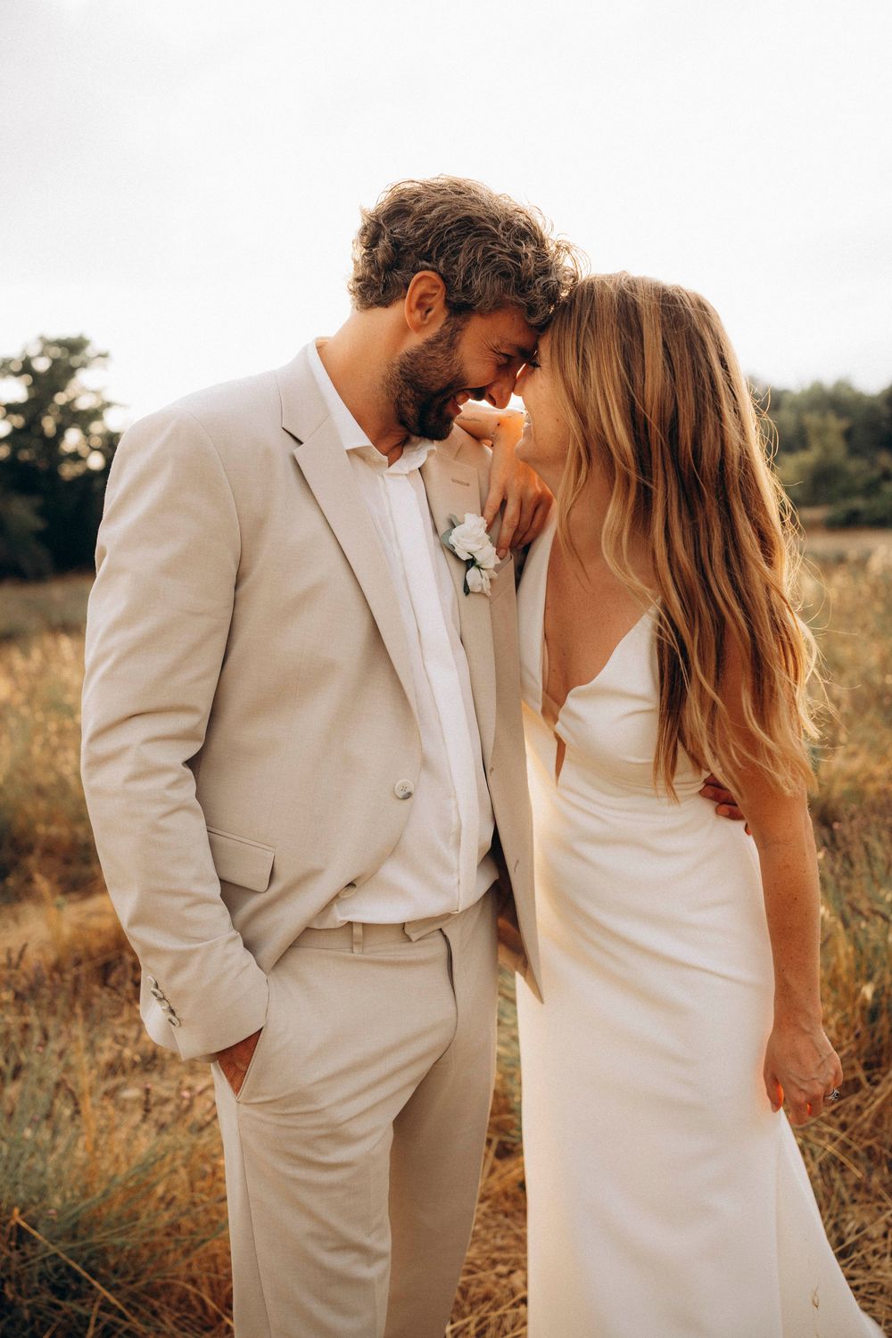 Bride and groom share an intimate moment in a golden field, wearing white dress and beige suit with floral boutonniere.