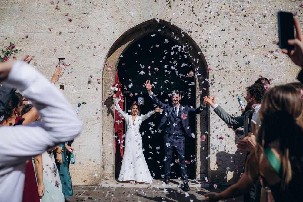 Newlyweds celebrate with confetti outside a stone church arch, surrounded by cheering guests at a joyful wedding ceremony.