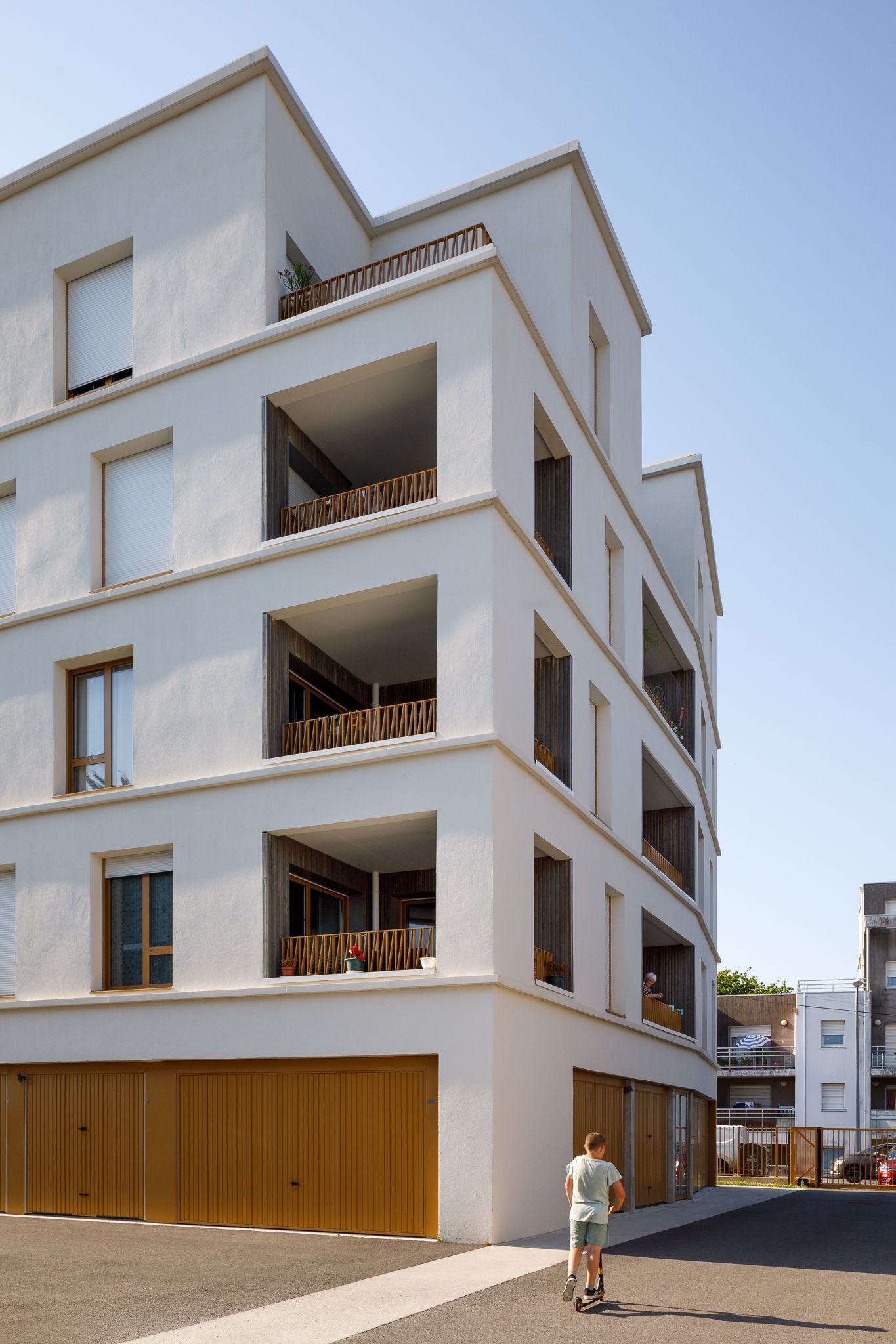 Immeuble d'appartements contemporain blanc avec balcons et portes de garage en bois ; une personne se tient au premier plan.