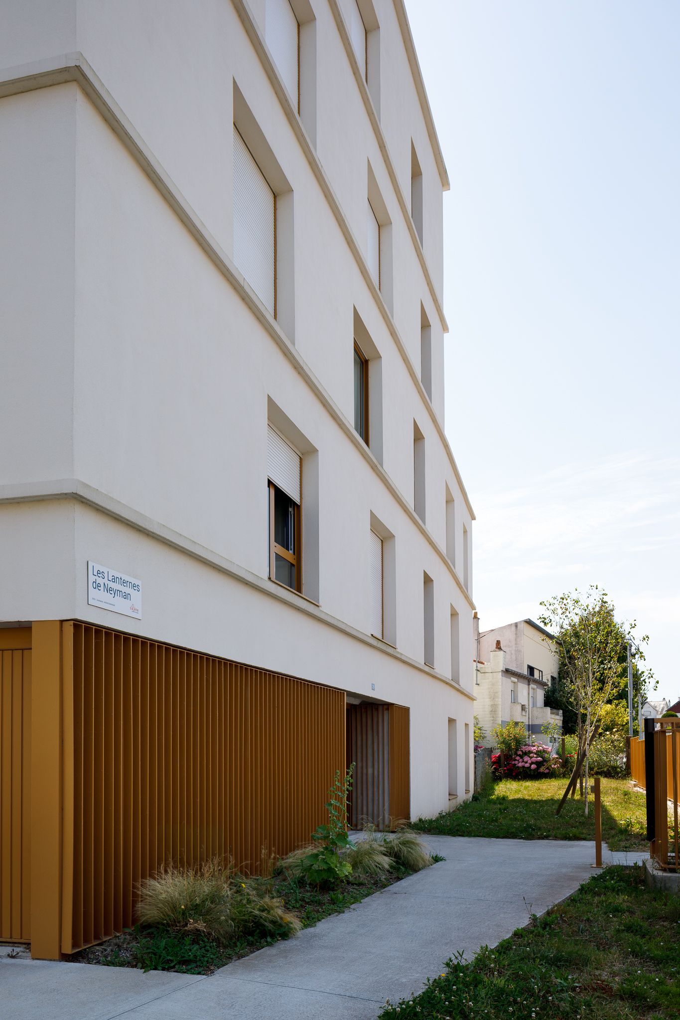 Immeuble d'appartements moderne à enduit crème, doté d'un bardage métallique jaune doré à la base et bordé d'une allée paysagère.