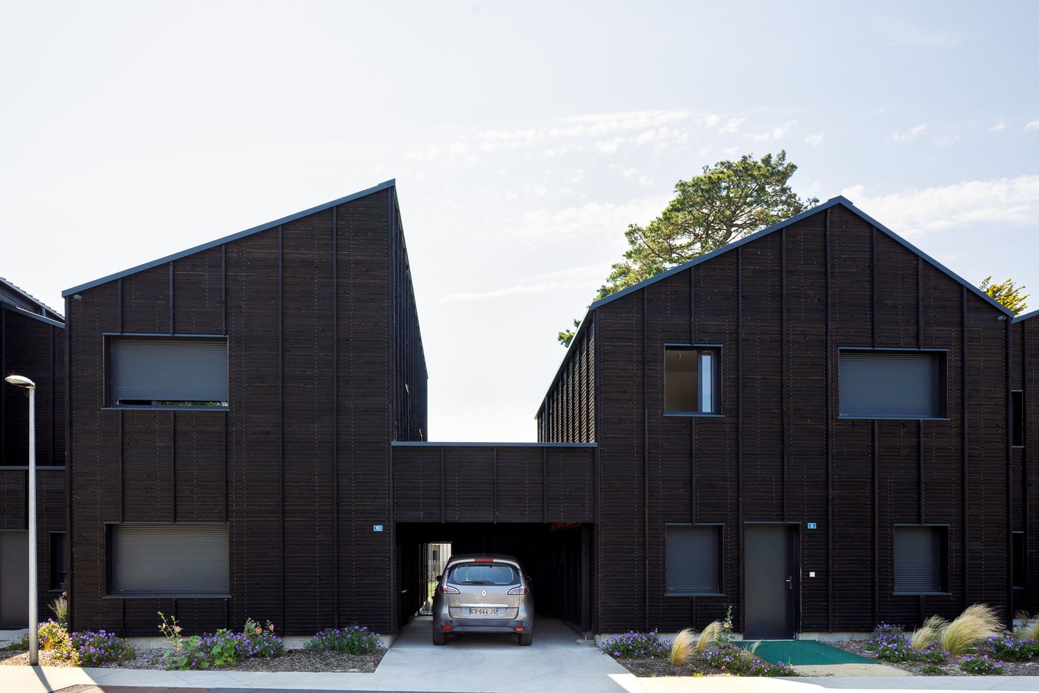 Maisons jumelées revêtues de bois gris anthracite, dotées de toits à pignons abrupts et d'un abri voiture, sous un ciel lumineux.