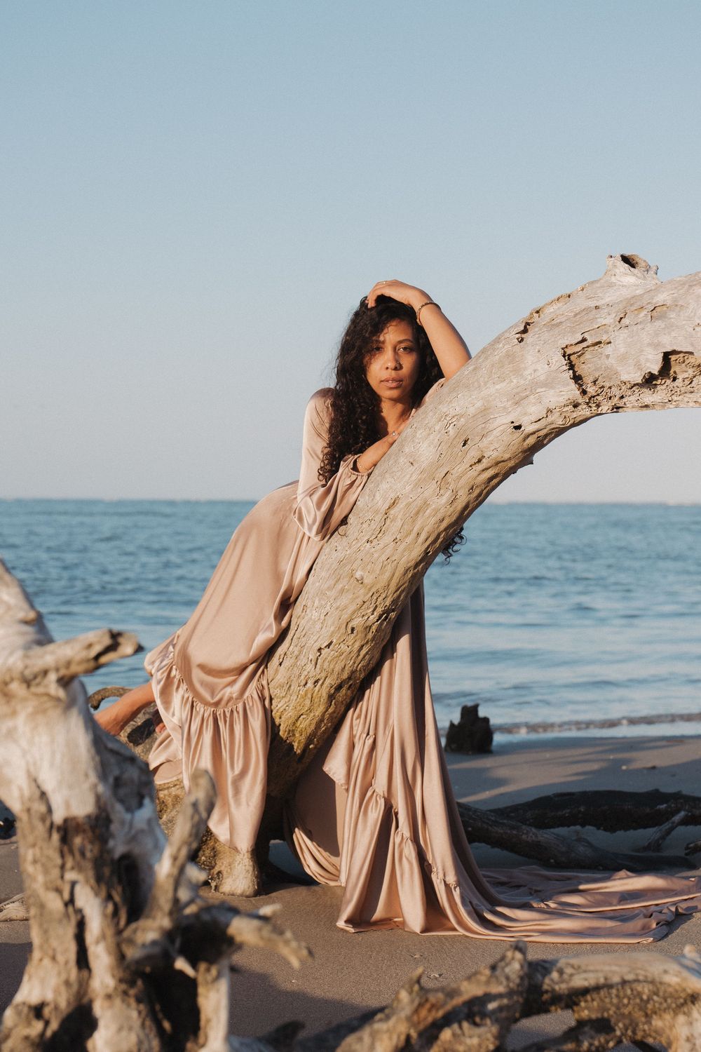 Woman in flowing beige gown poses against driftwood on a serene beach at golden hour with ocean backdrop