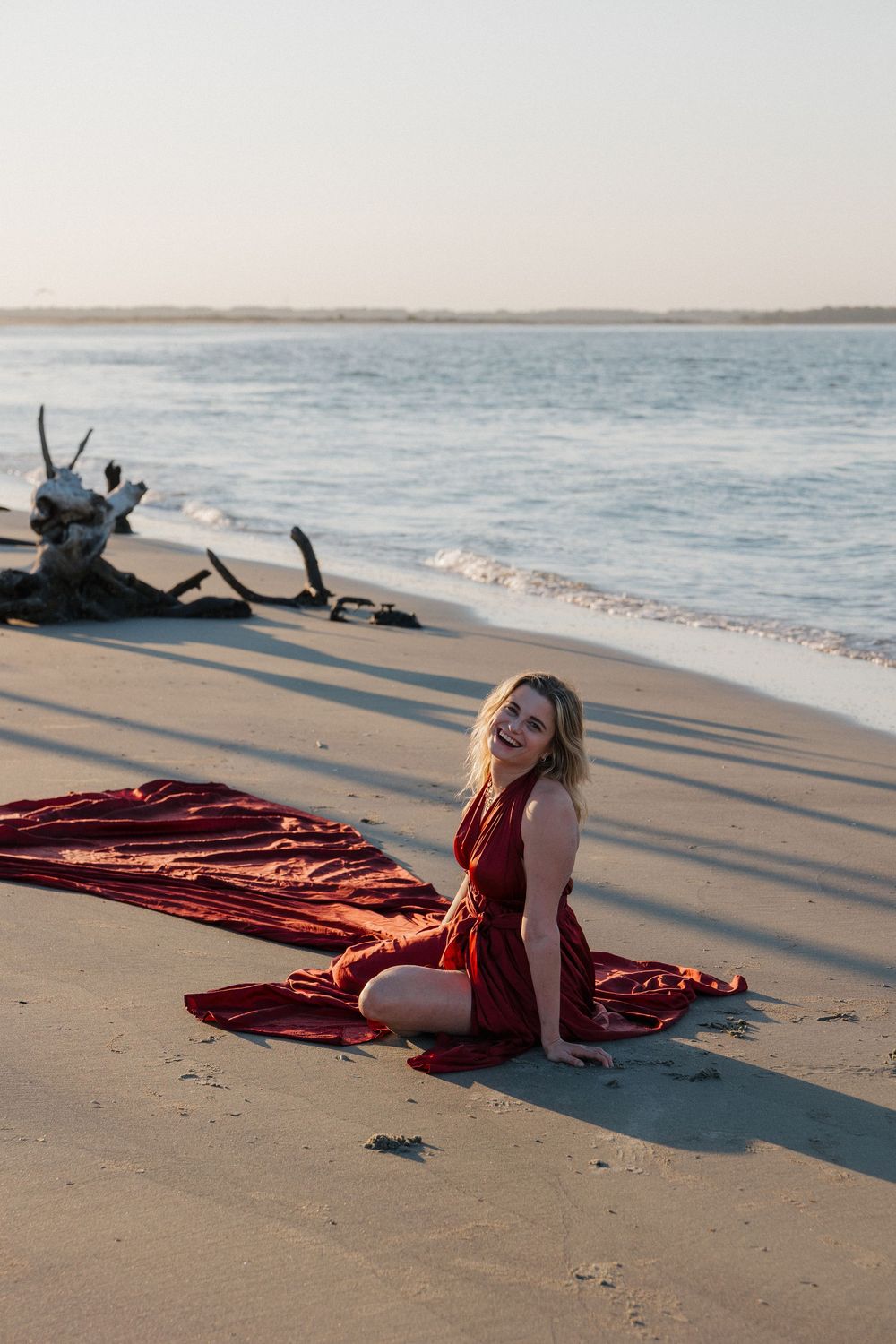 Woman in red dress sitting on sandy beach at sunset with driftwood and calm ocean waves behind her.