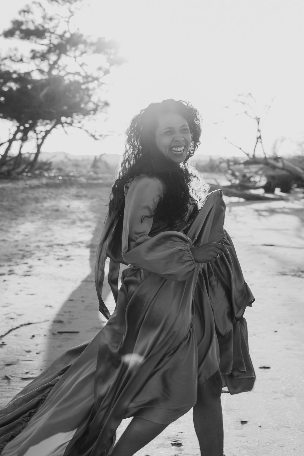 Black and white photo of laughing woman in flowing satin dress walking on sunlit sandy beach.