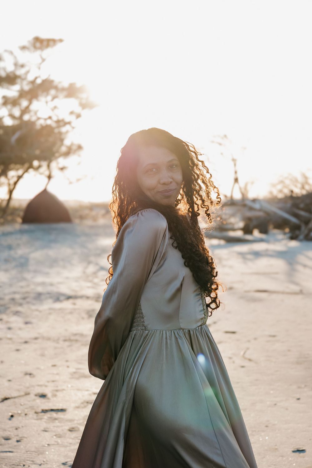 Woman in stone colored off-shoulder gown with curly hair smiling over shoulder at golden hour beach.