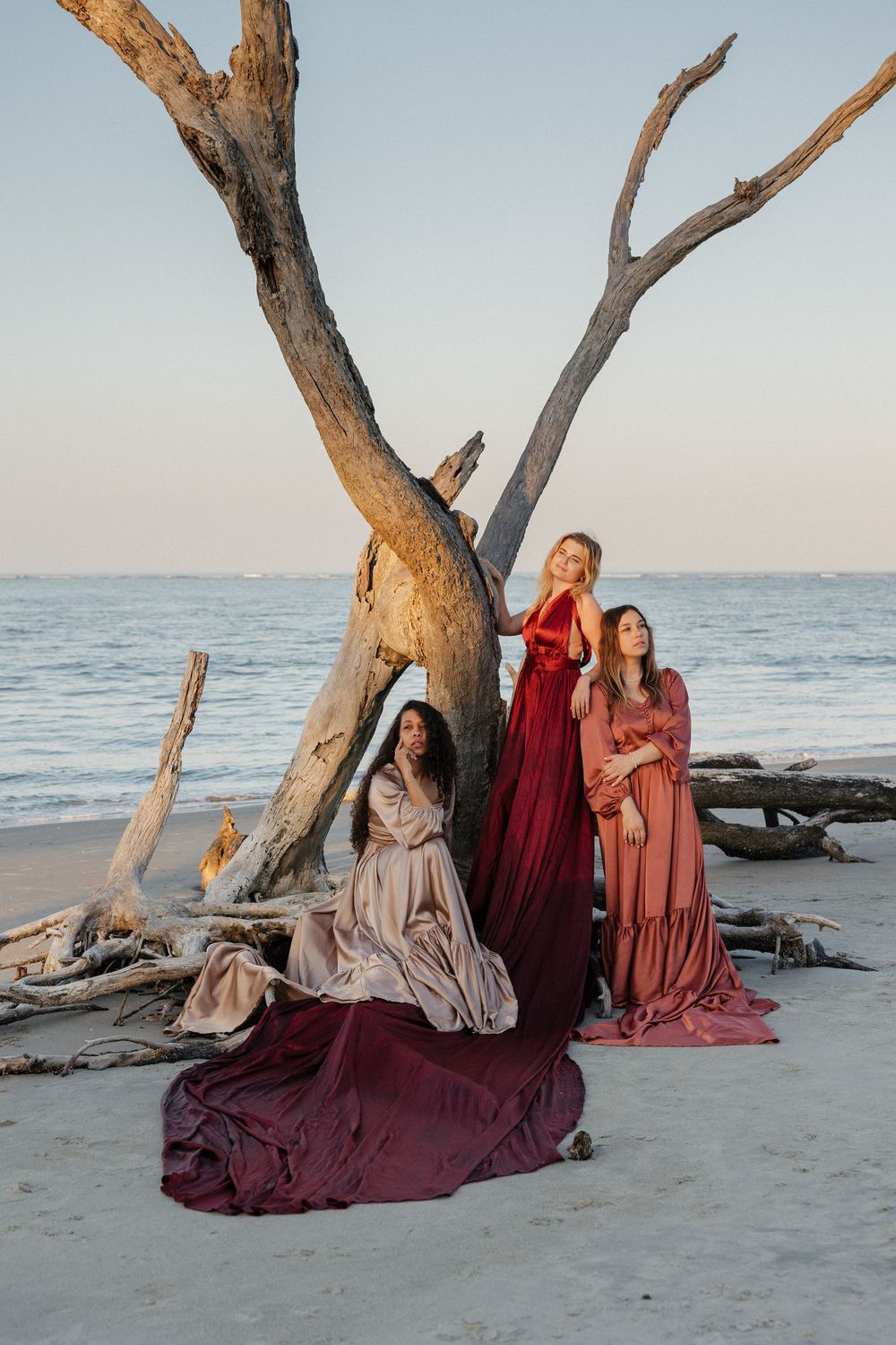 Three women in elegant flowing gowns pose by driftwood on a beach at sunset.
