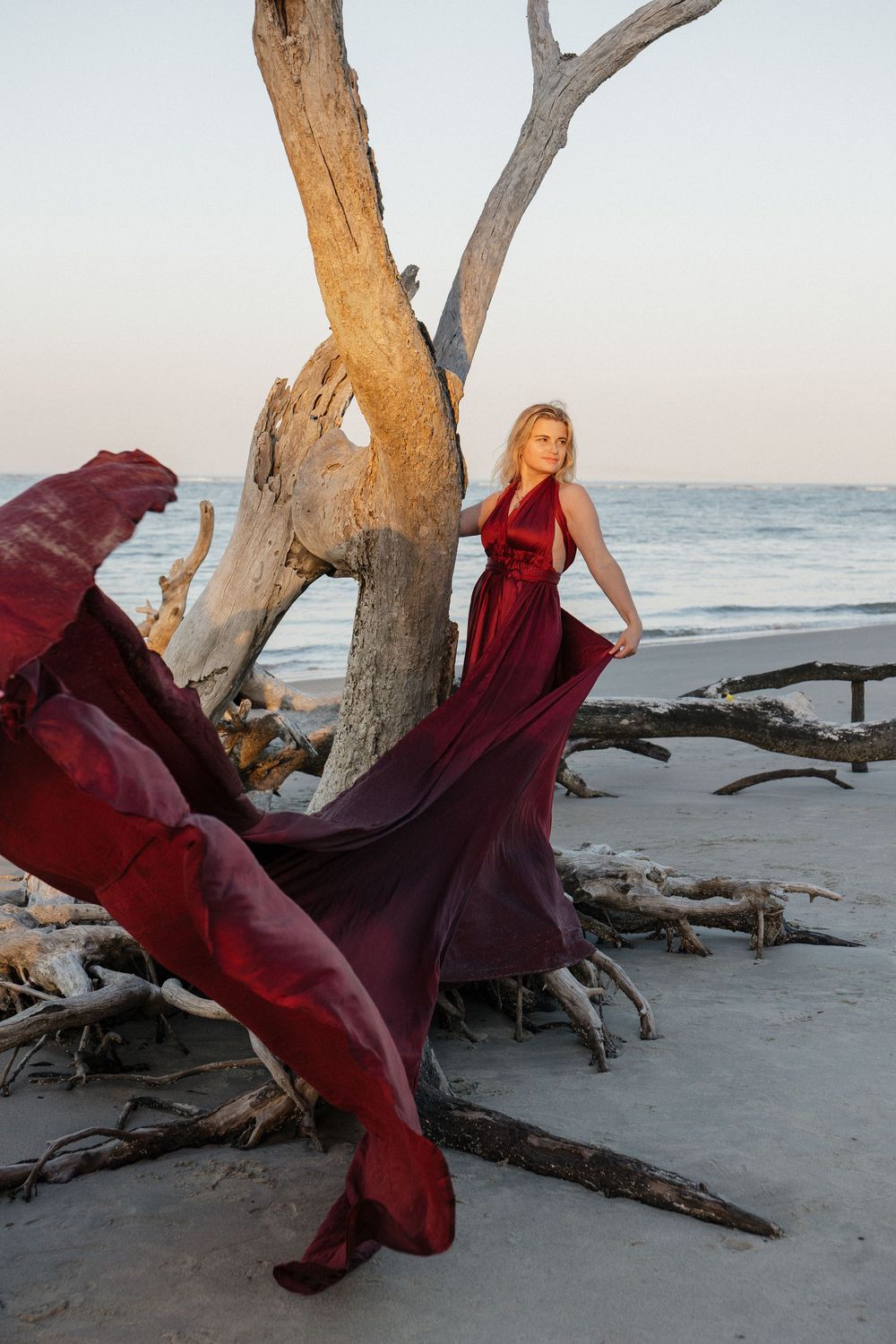Blonde woman in flowing burgundy gown posing beside driftwood tree on serene ocean beach at sunset.