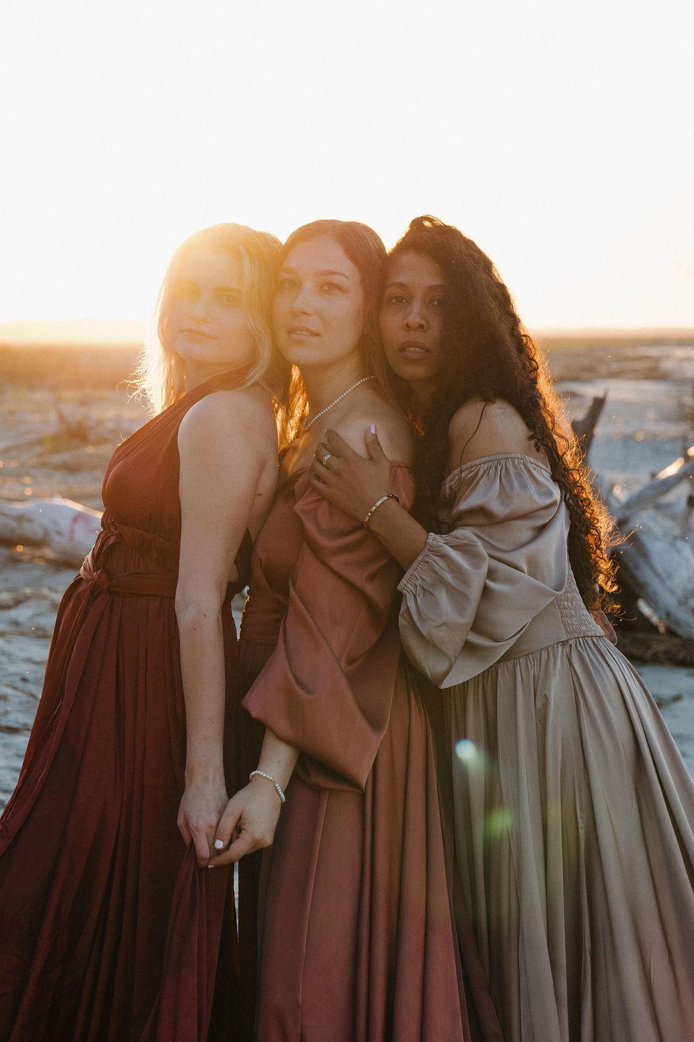 Three women in elegant rust, burgundy, and taupe gowns pose together at golden hour on a sandy beach.