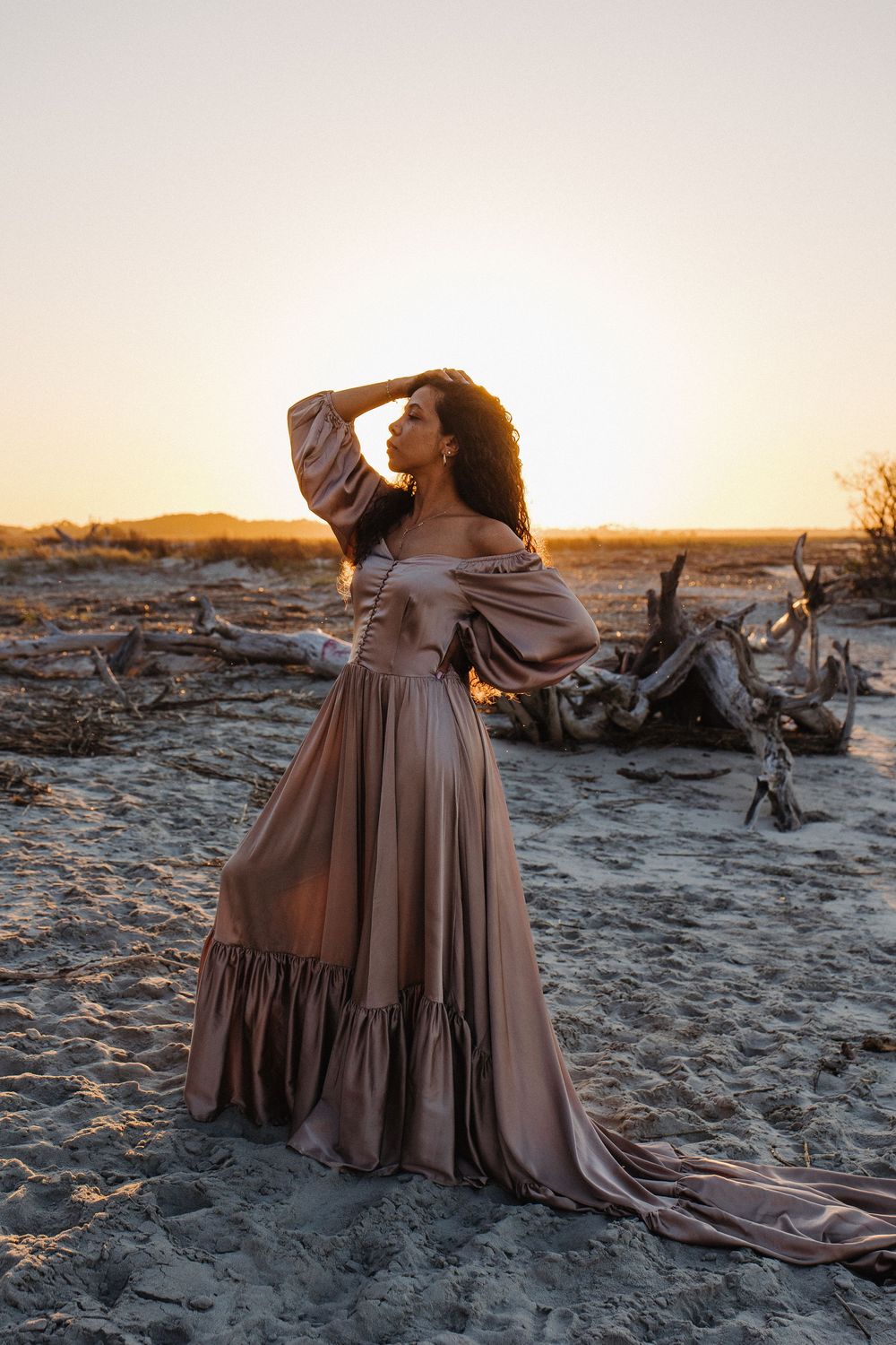 Woman in flowing champagne off-shoulder gown poses gracefully at sunset on a sandy desert beach.