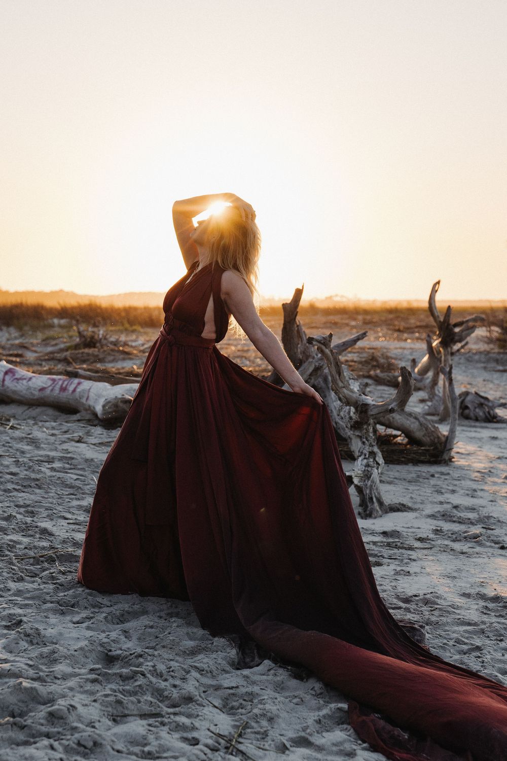 Woman in flowing dark red gown poses dramatically on sandy beach at golden sunset near driftwood.