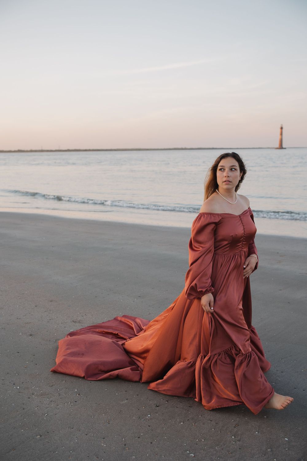 Woman in rust satin off-shoulder gown with train walks barefoot on beach at dusk near lighthouse.