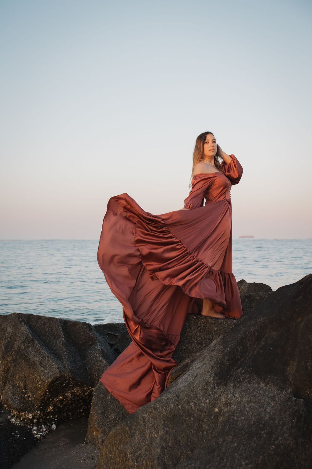 Woman in flowing rust-colored satin gown standing on rocky shore at sunset with ocean backdrop.