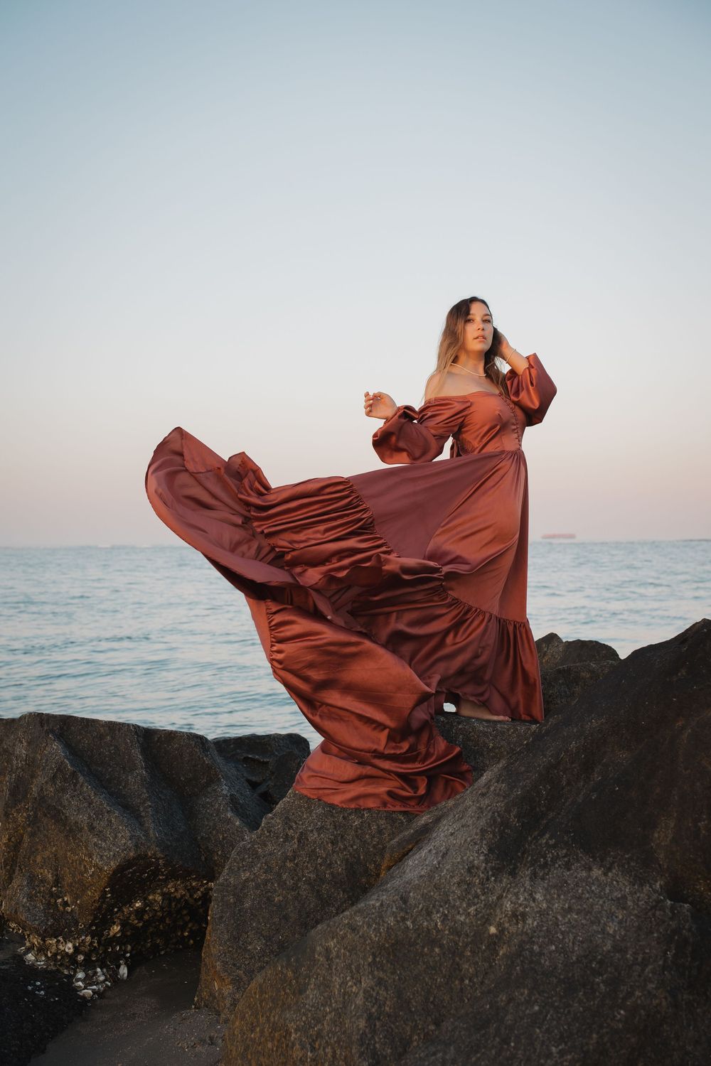 Woman in billowing rust satin gown stands on rocky shore at sunset, dress flowing in the wind.