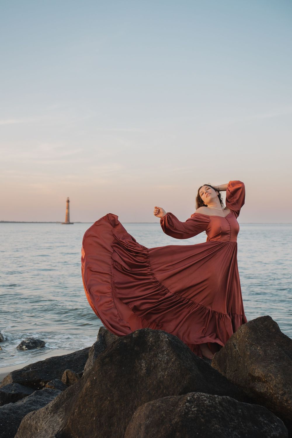Woman in flowing rust gown dances on coastal rocks at dusk with lighthouse visible in background.
