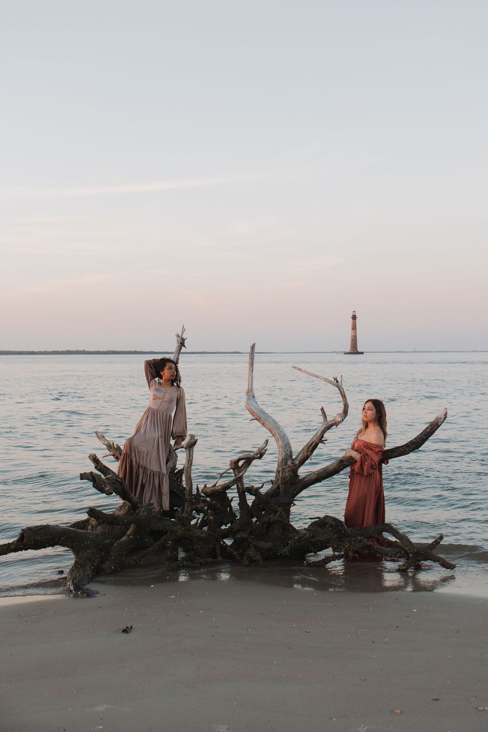 Two women in elegant gowns pose on driftwood at beach sunset with lighthouse in the distance.