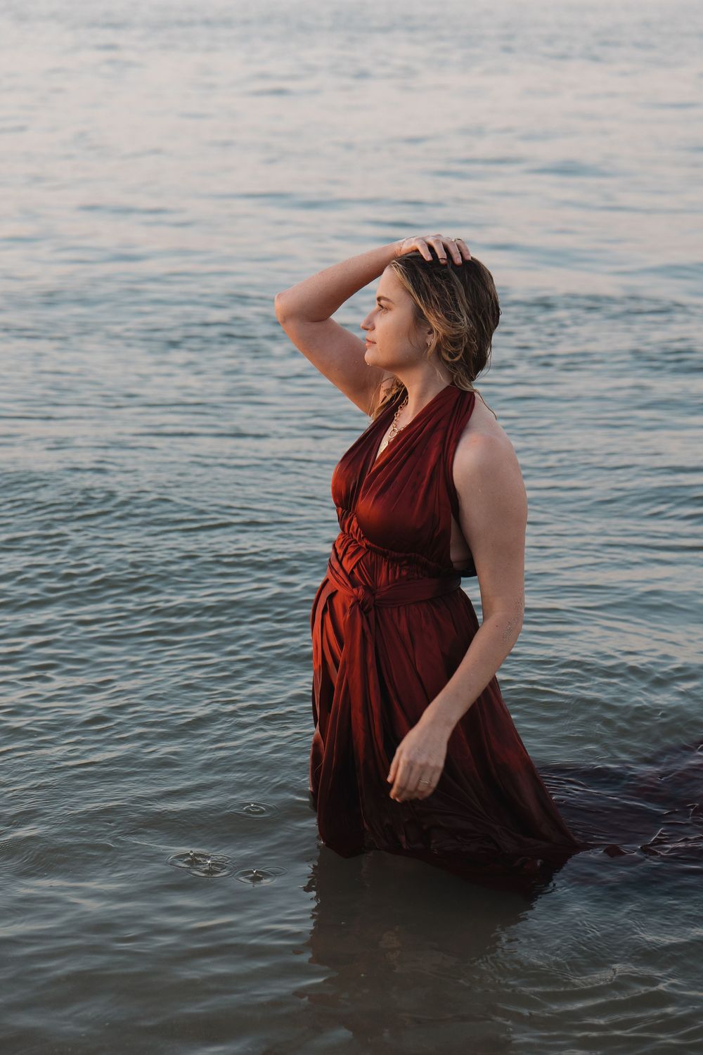 Woman in deep red halter gown stands waist-deep in calm ocean water at dusk, hand in hair.