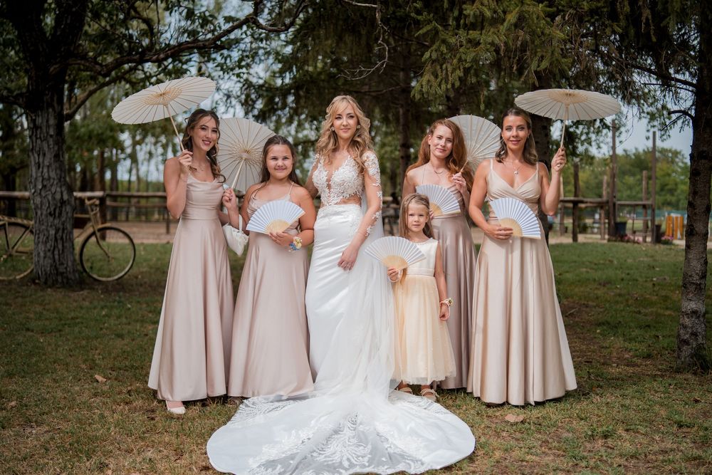Bride in white gown poses with bridesmaids in champagne dresses holding parasols outdoors at rustic wedding venue.