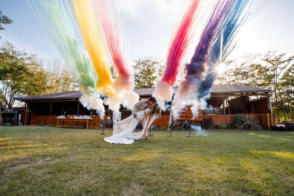 Couple shares romantic dip kiss surrounded by colorful smoke bomb rainbow at outdoor wedding celebration.