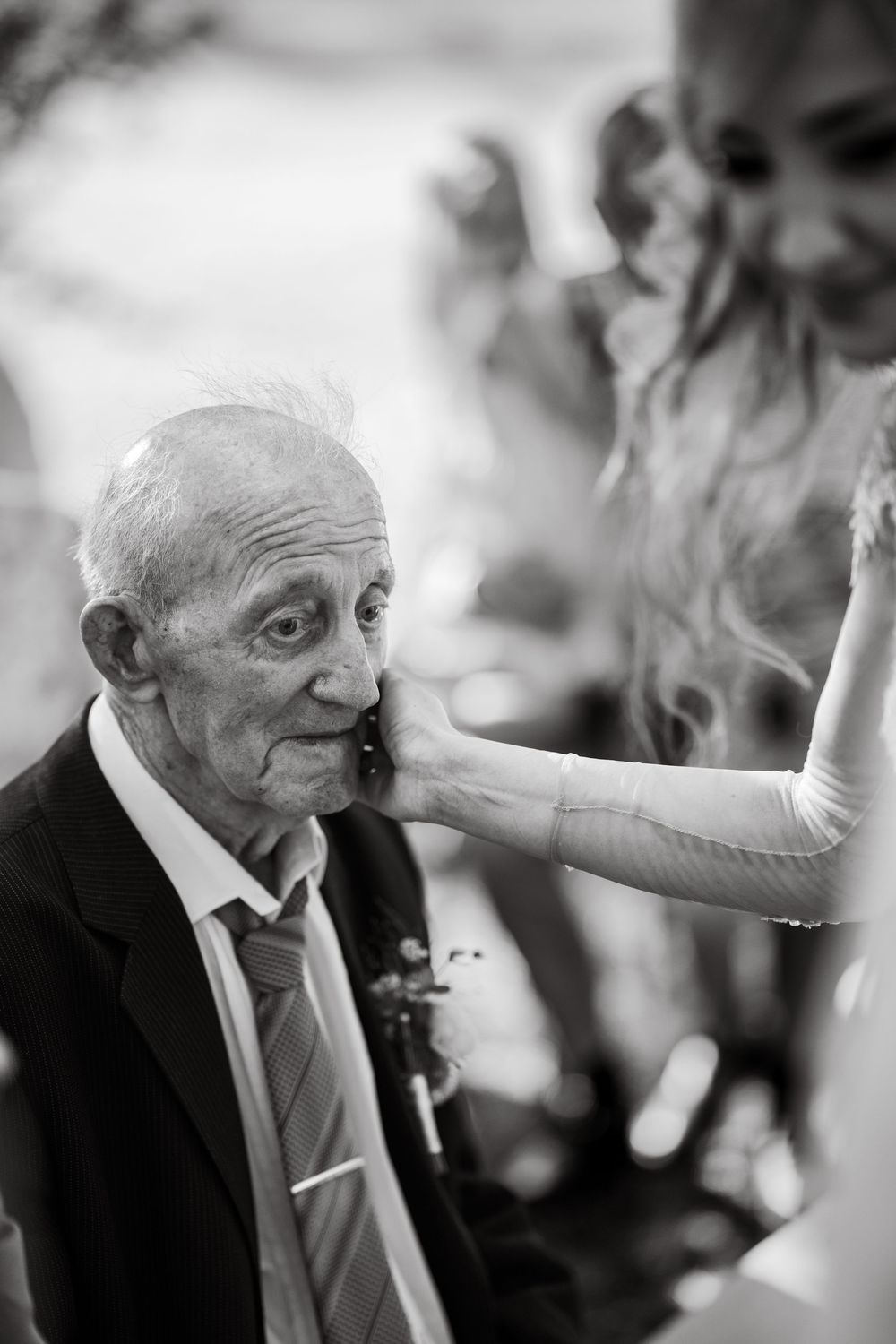 Black and white photo of elderly man in suit and tie having his cheek touched tenderly at a wedding.