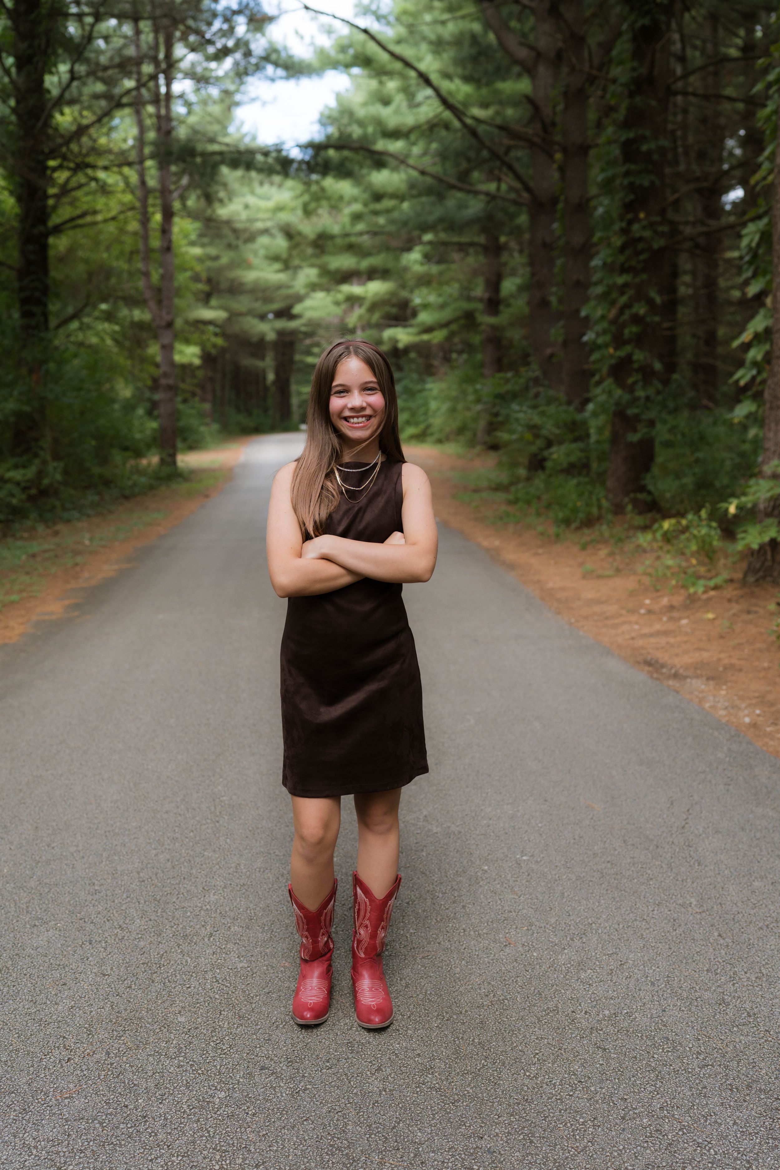 Young woman in black dress and red cowboy boots stands smiling on a tree-lined road with arms crossed.