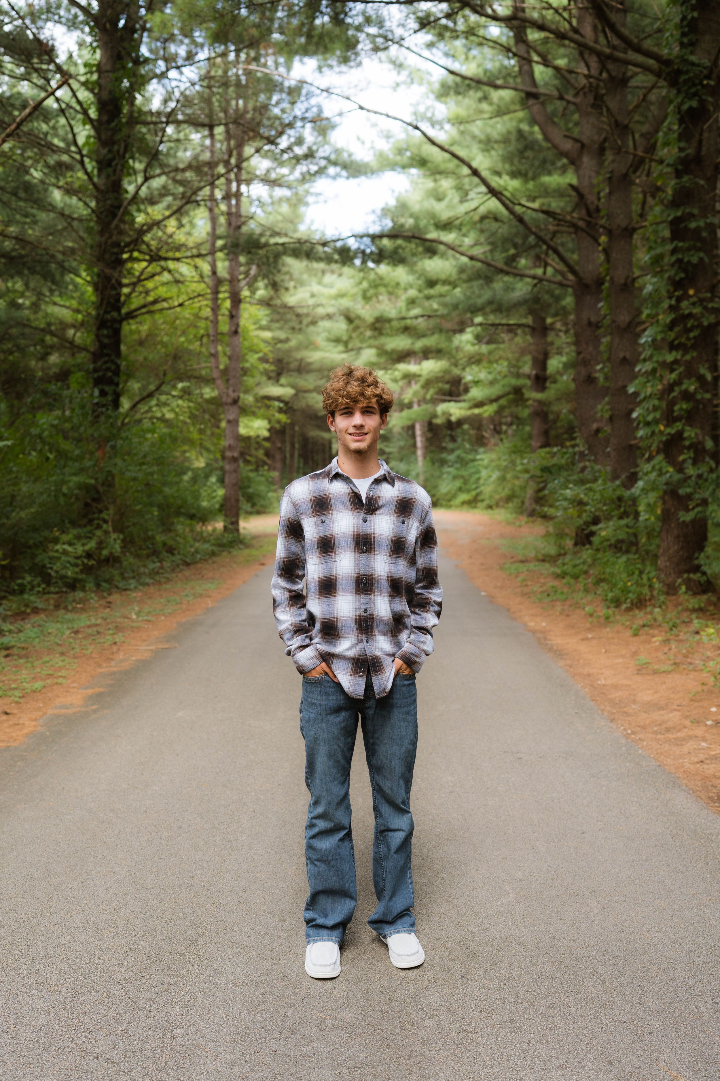 Teen boy with curly hair in plaid shirt and jeans standing on a tree-lined road in a forest setting.