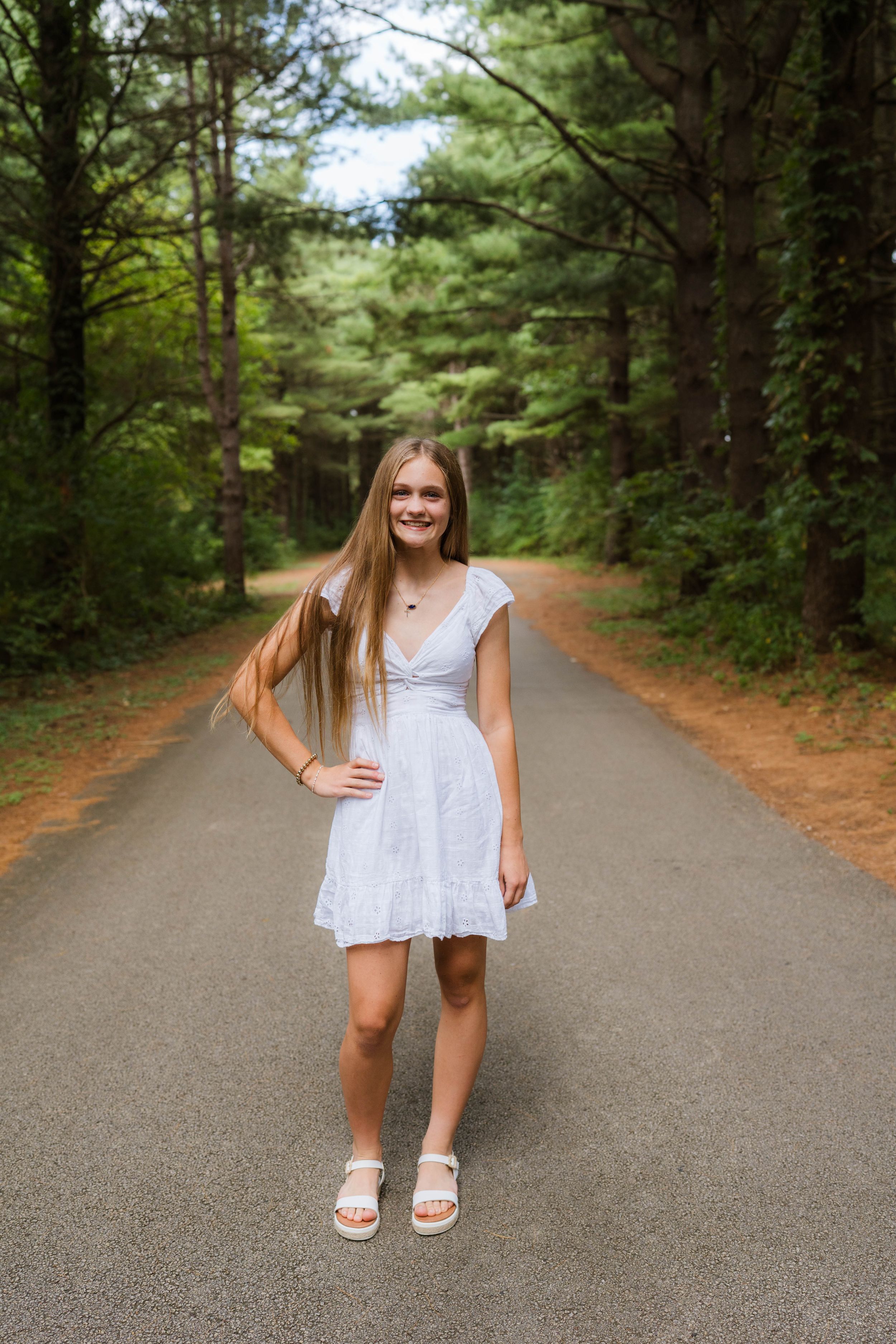 Young woman in white dress standing on tree-lined road, smiling confidently with long blonde hair and white sandals.