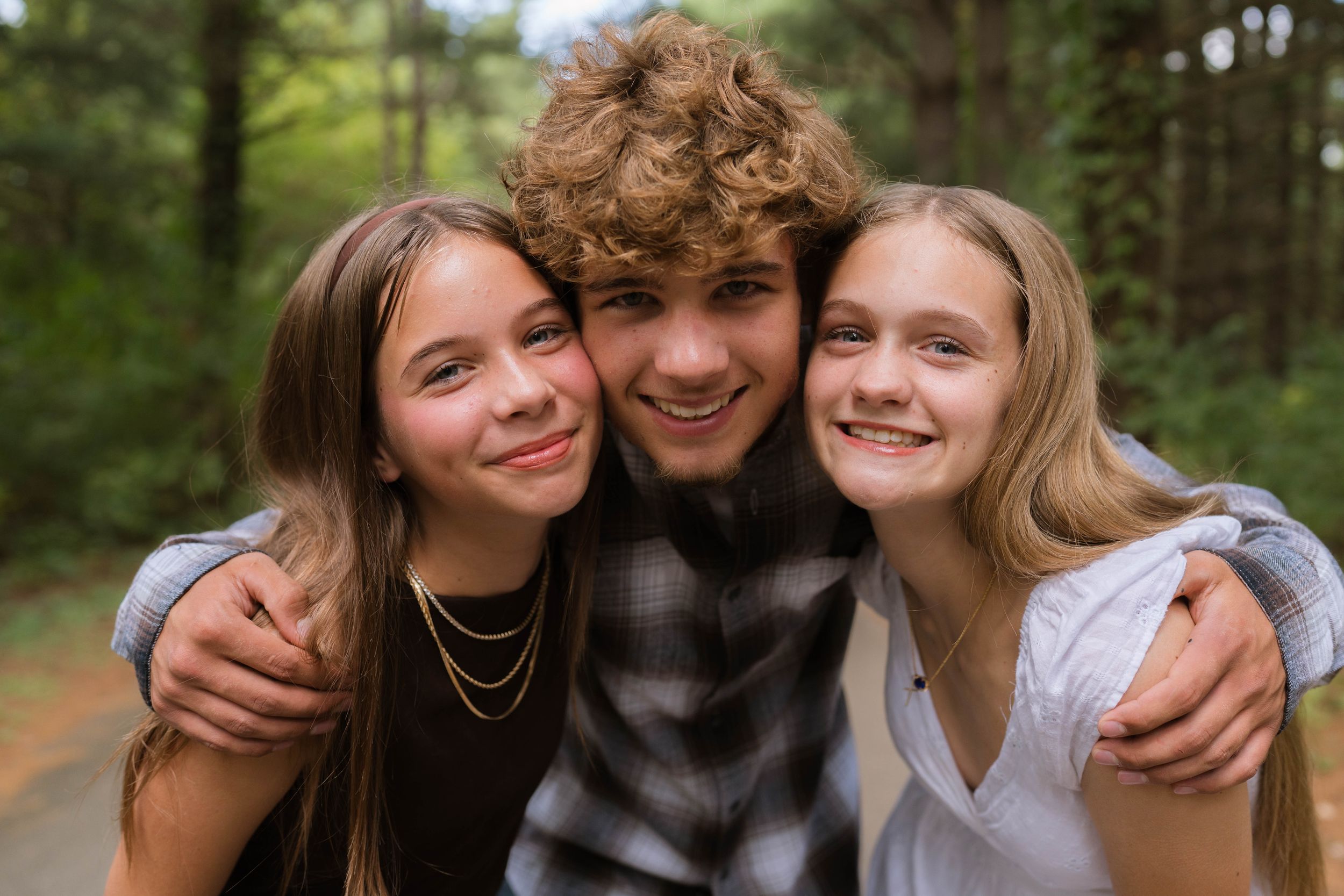 Three smiling teenagers pose together on a forest path, with a boy in a plaid shirt between two girls.