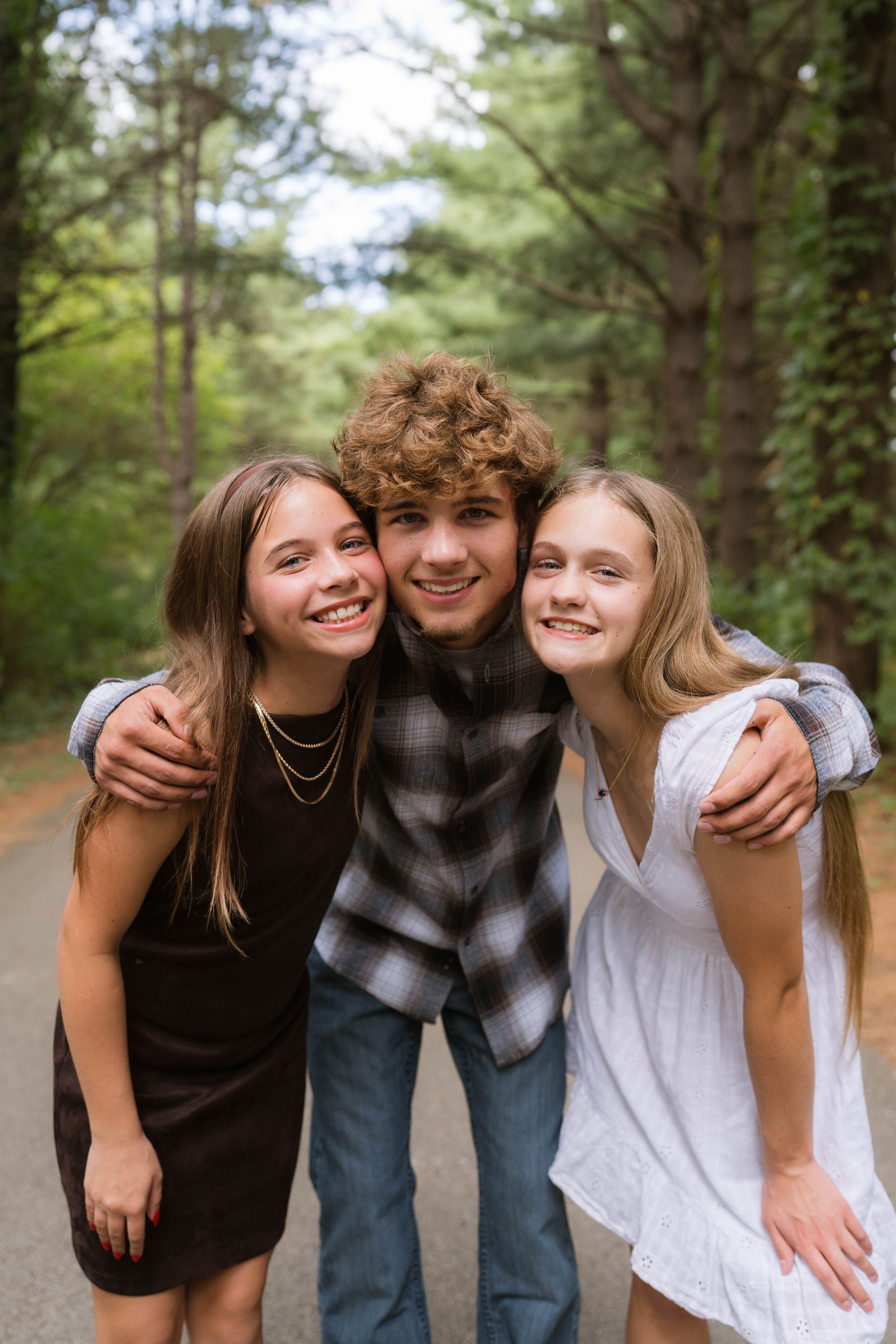Three smiling teenagers posing together on a tree-lined path, two girls flanking a curly-haired boy.