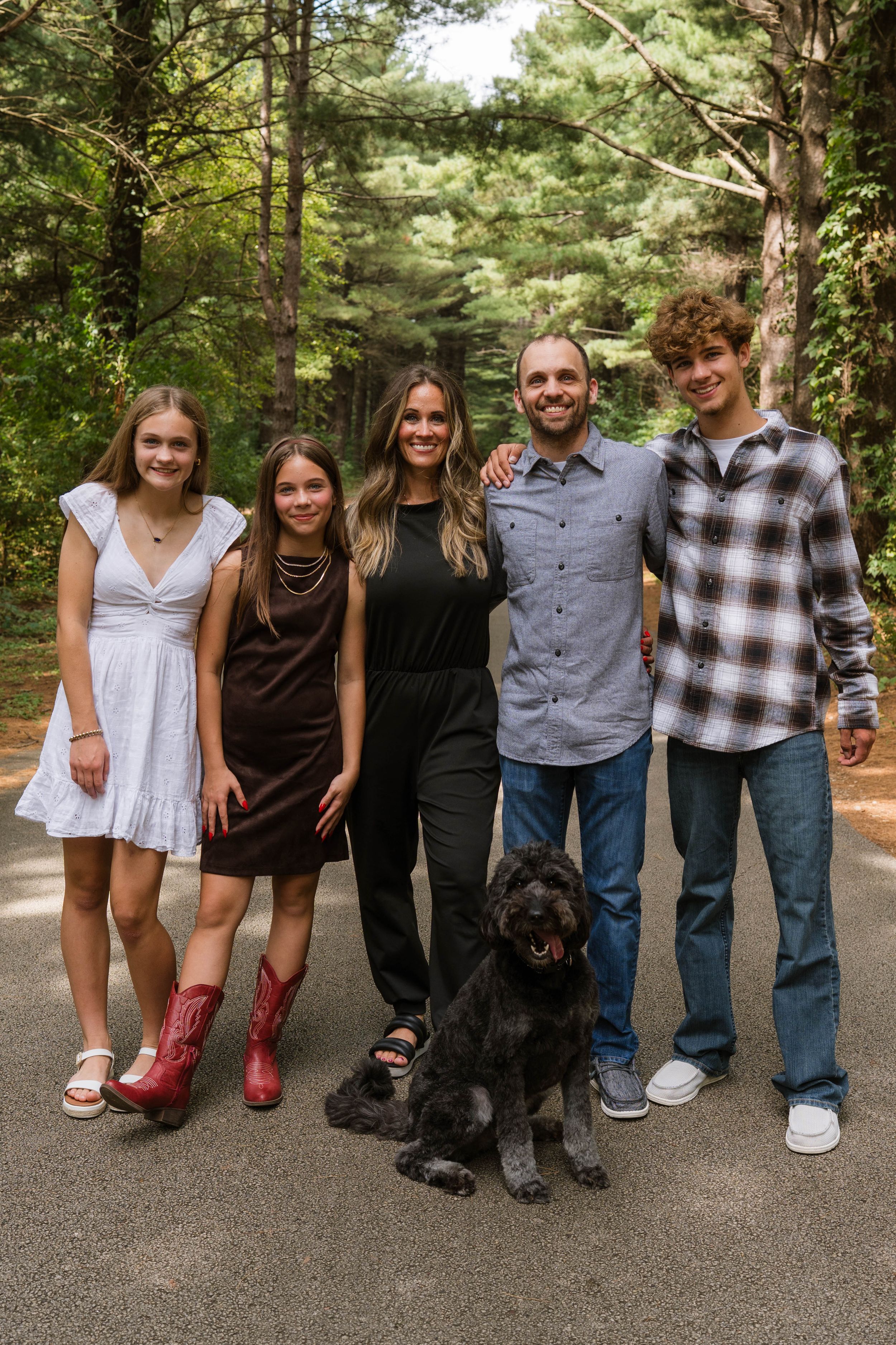 Family of five poses with black dog on wooded path, wearing casual attire including plaid shirt and cowboy boots.