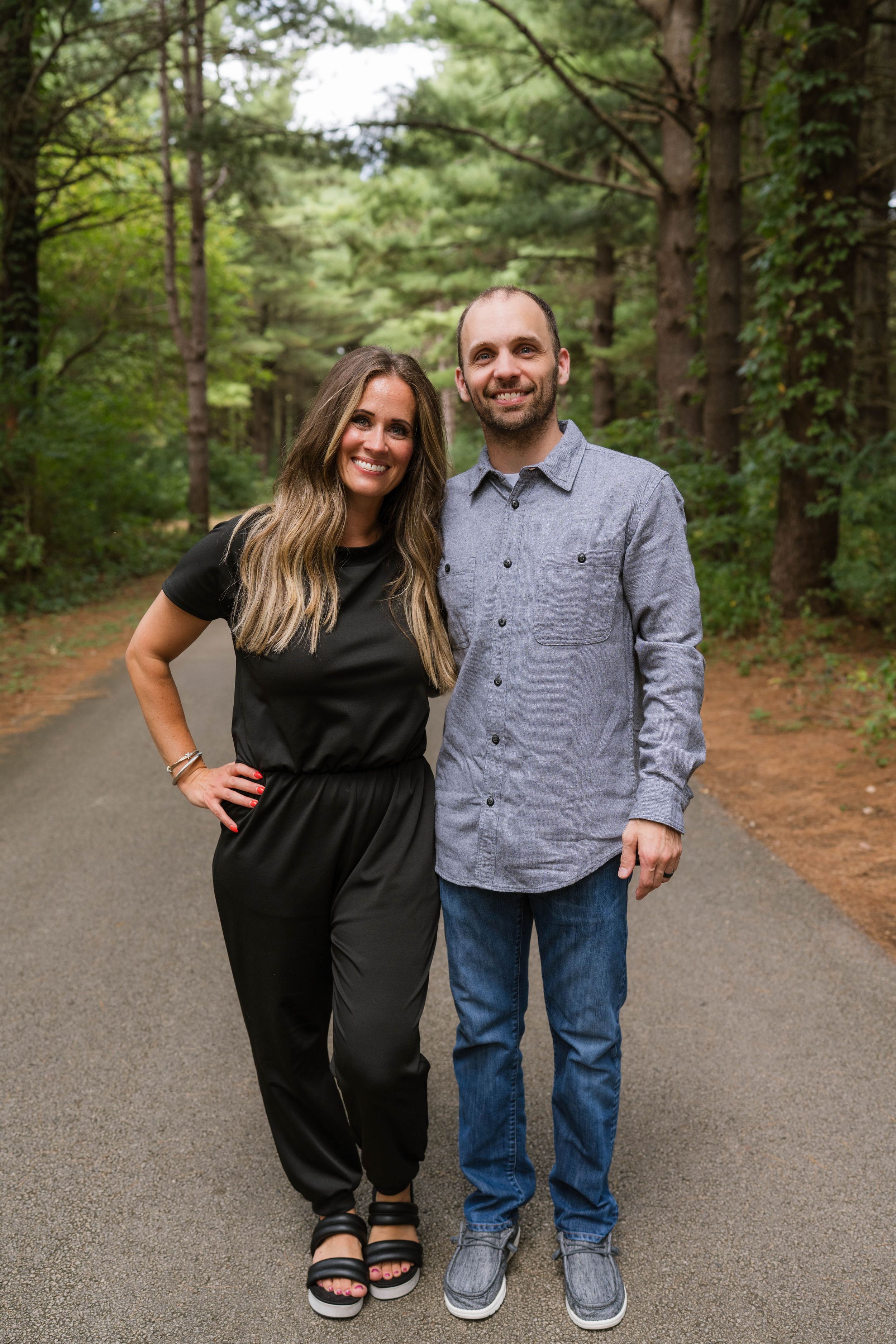 A smiling couple poses on a tree-lined path, woman in black jumpsuit and man in gray shirt with jeans.